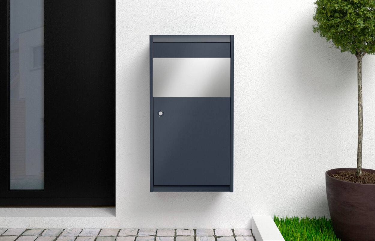 Une boîte aux lettres moderne en gris foncé est suspendue à un mur blanc à côté d'une grande porte. Un arbre en pot se trouve à proximité.