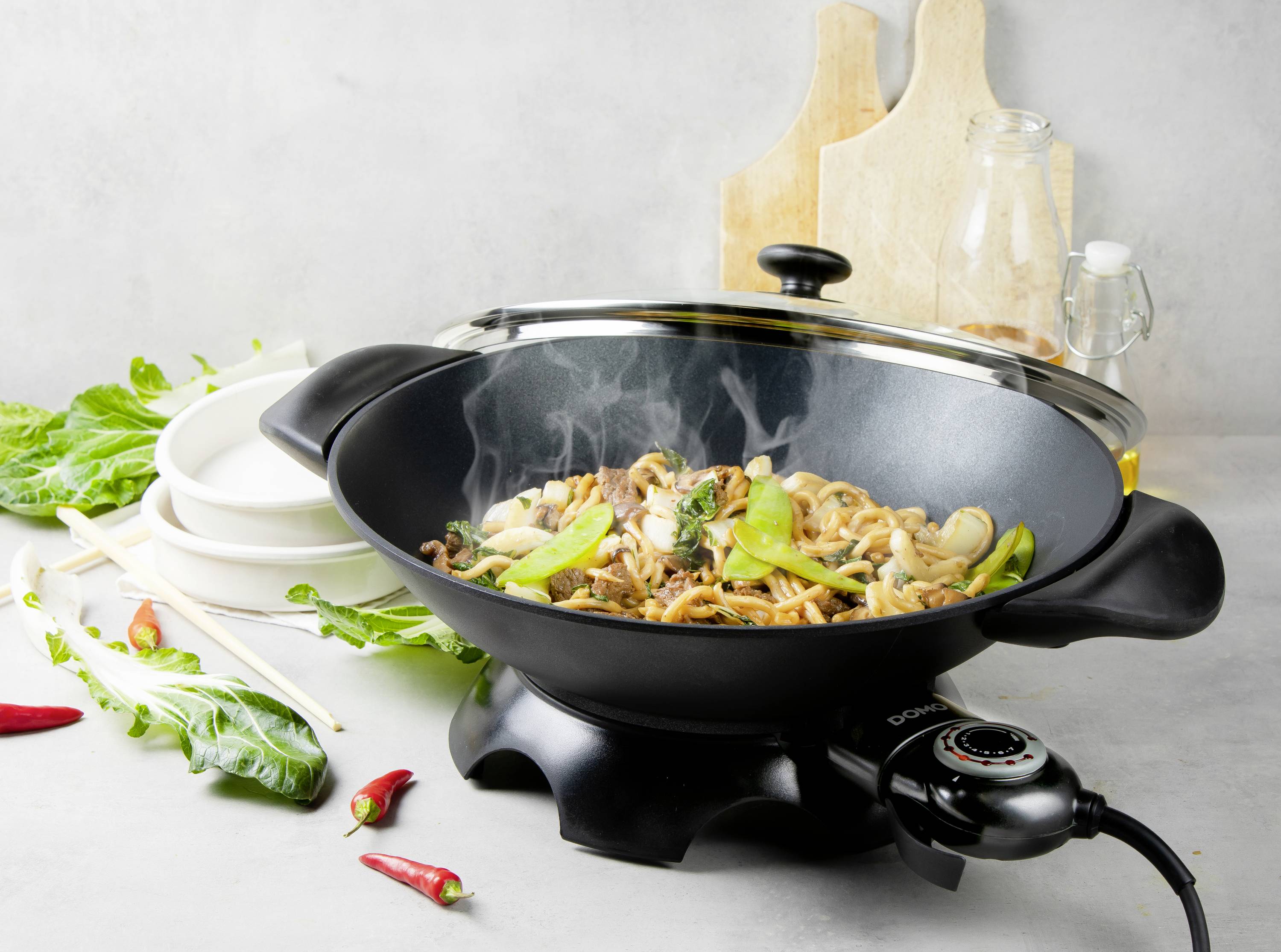 Un wok avec un sauté de légumes asiatiques fumant, composé de nouilles, de légumes verts et de champignons. En arrière-plan, des épices et des assiettes.
