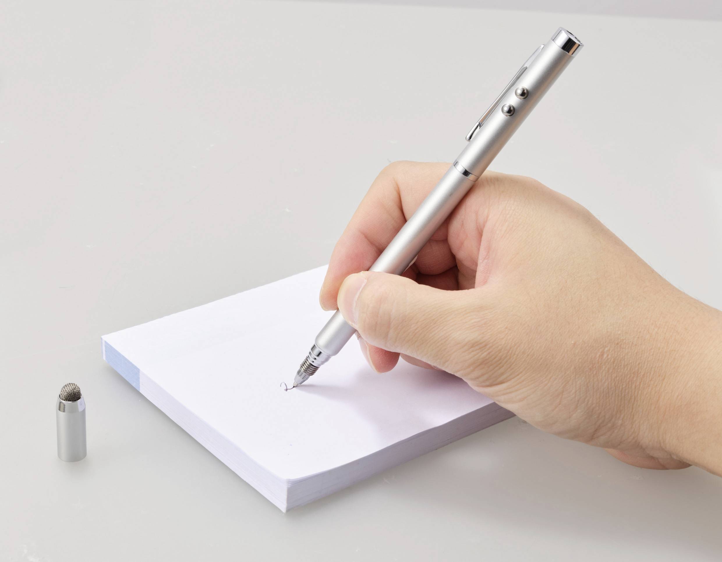 Une main écrit avec un stylo argenté sur un bloc-notes. Le capuchon du stylo est posé à côté.