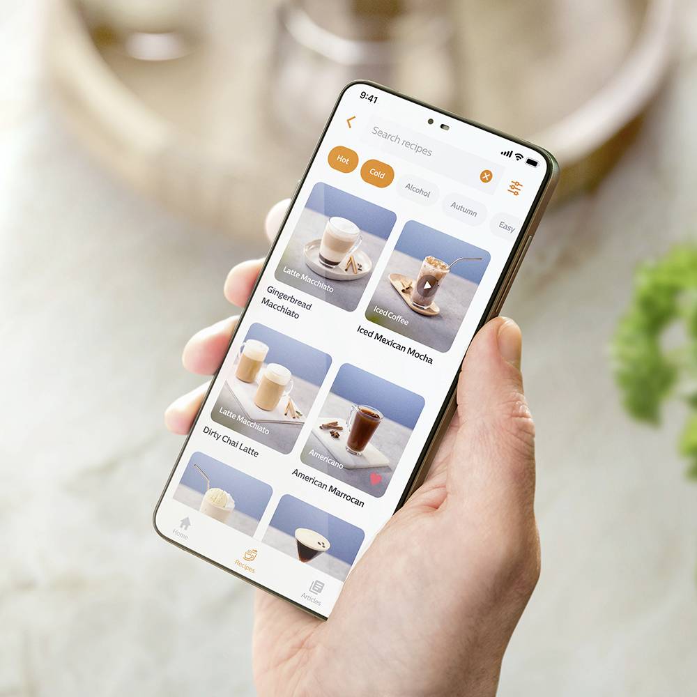 Une main tient un smartphone affichant une sélection de boissons dans une application, incluant un Café Latte, un Café et des Milkshakes.