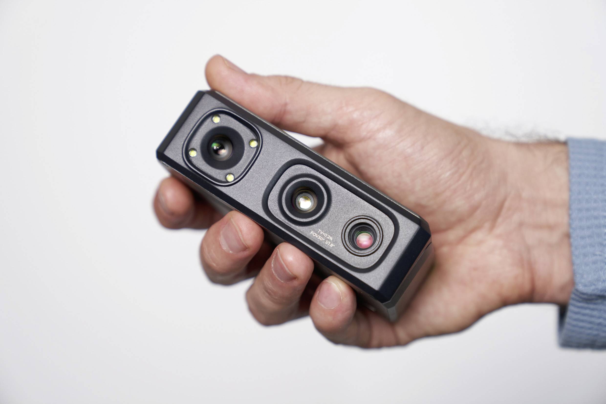 Une main tient un petit appareil photo rectangulaire doté de plusieurs objectifs et capteurs. L'appareil est noir et présente une structure compacte.