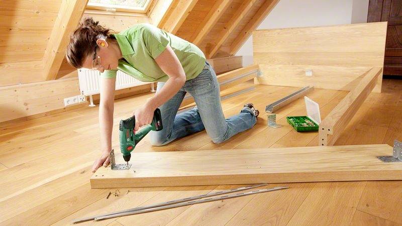 Une femme monte une étagère en bois dans le grenier. Elle porte des protections auditives et utilise un visseuse sans fil. Autour d'elle sont disposés des outils et des planches.