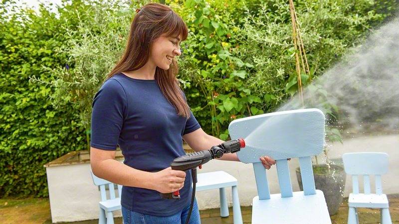 Une femme nettoie une chaise bleue en bois avec un nettoyeur haute pression dans le jardin. À l'arrière-plan, on aperçoit d'autres chaises bleues et des plantes.