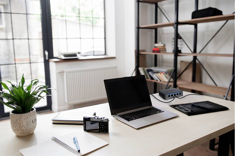Un bureau moderne avec un ordinateur portable, une tablette graphique, un appareil photo, un carnet de notes et une plante en pot. En arrière-plan, une grande fenêtre et une bibliothèque.