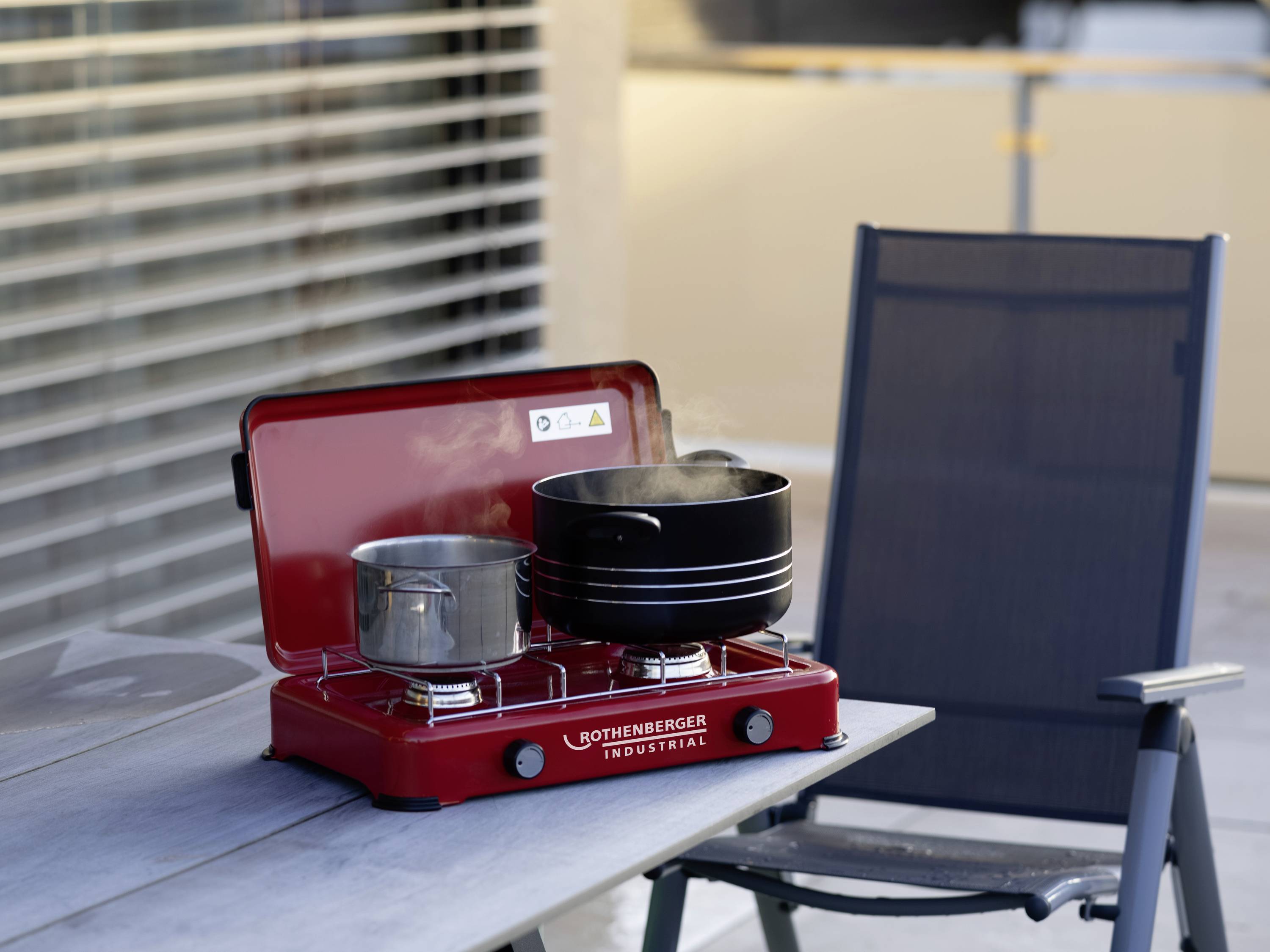 Un réchaud à gaz rouge sur une table à l'extérieur, avec deux casseroles d'où s'élève de la vapeur. À côté se trouve une chaise de jardin grise.