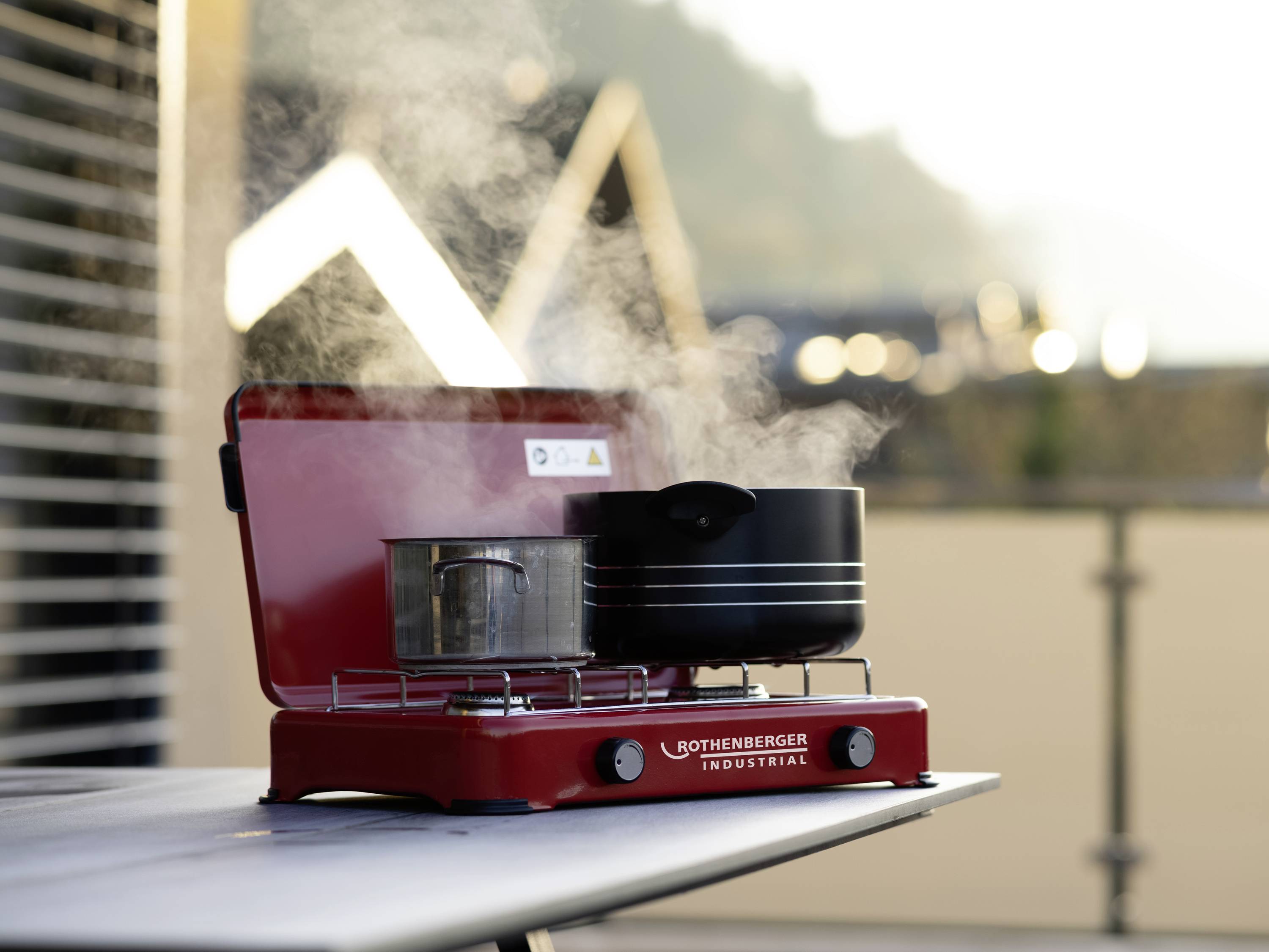 Un réchaud de camping rouge est posé à l'extérieur sur une table. Il est allumé et deux casseroles contenant des préparations fumantes sont disposées dessus.