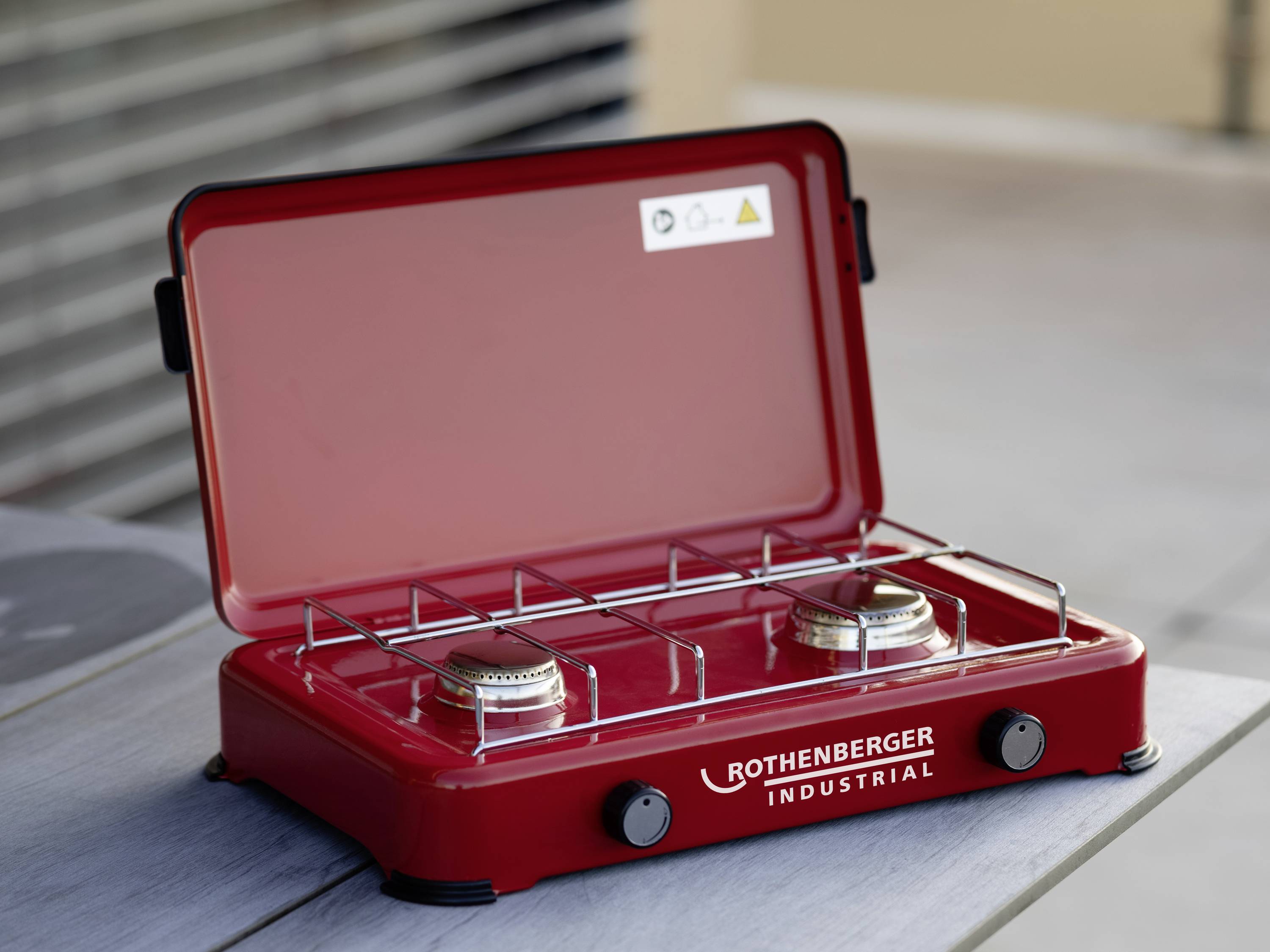Un réchaud à gaz portable rouge à deux brûleurs posé sur une table. En arrière-plan, flou, un dispositif de sécurité du bâtiment est visible sur le côté.