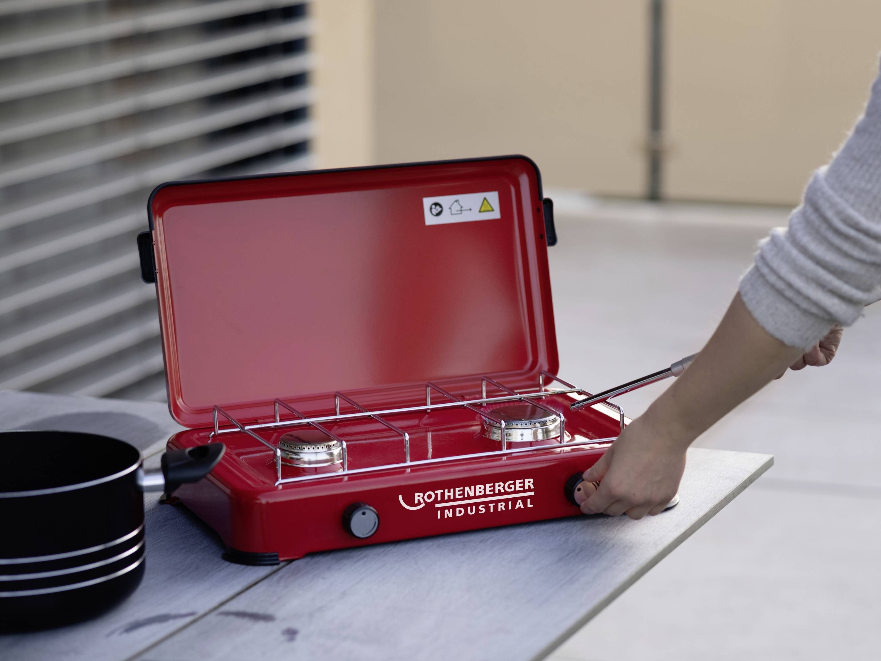 Une main place un réchaud à gaz portable rouge à deux brûleurs de 'Rothenberger Industrial' sur une table extérieure.