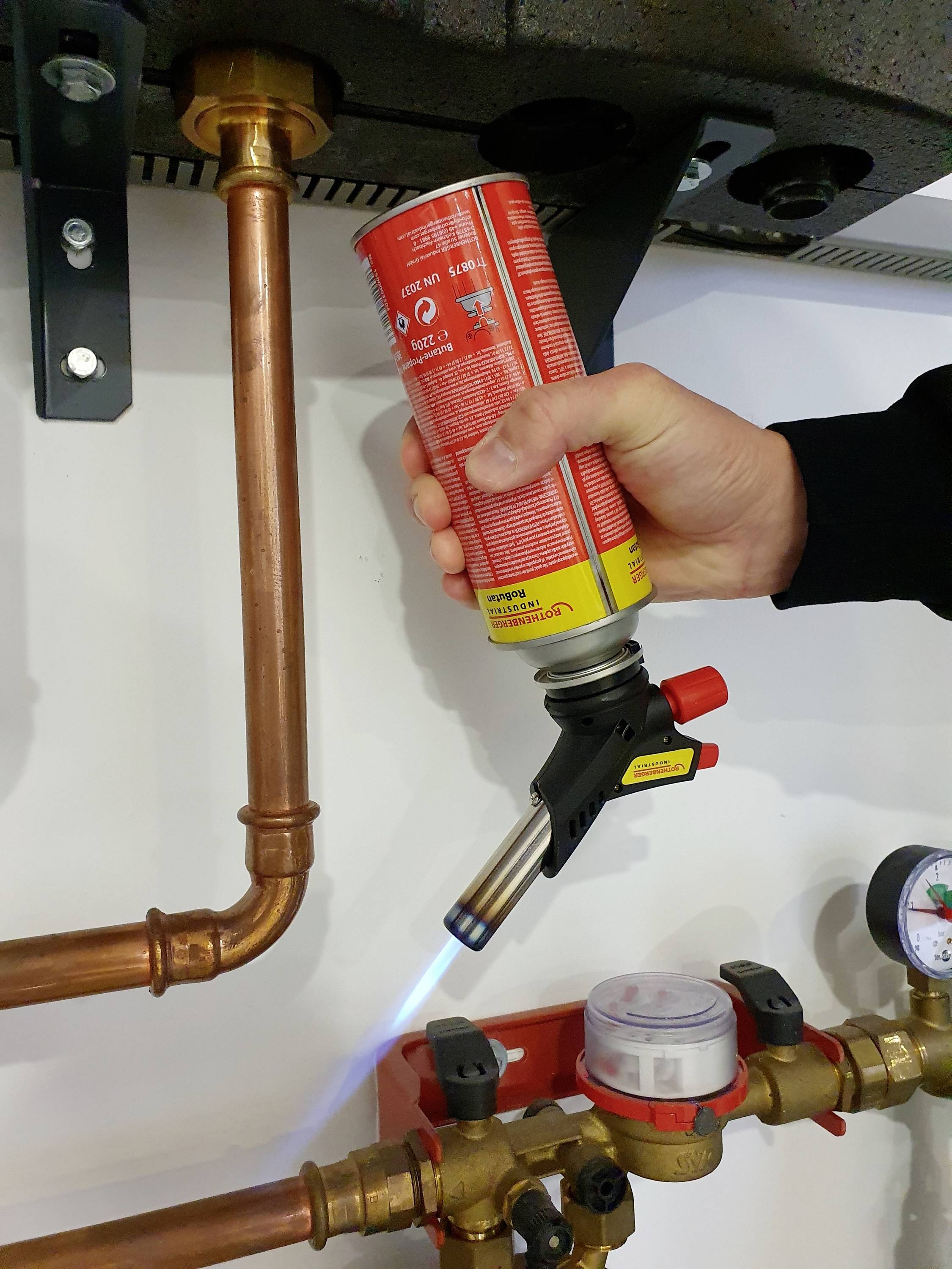 Une main tient un chalumeau et chauffe un tube en cuivre dans un système de chauffage. Un manomètre est visible en arrière-plan.