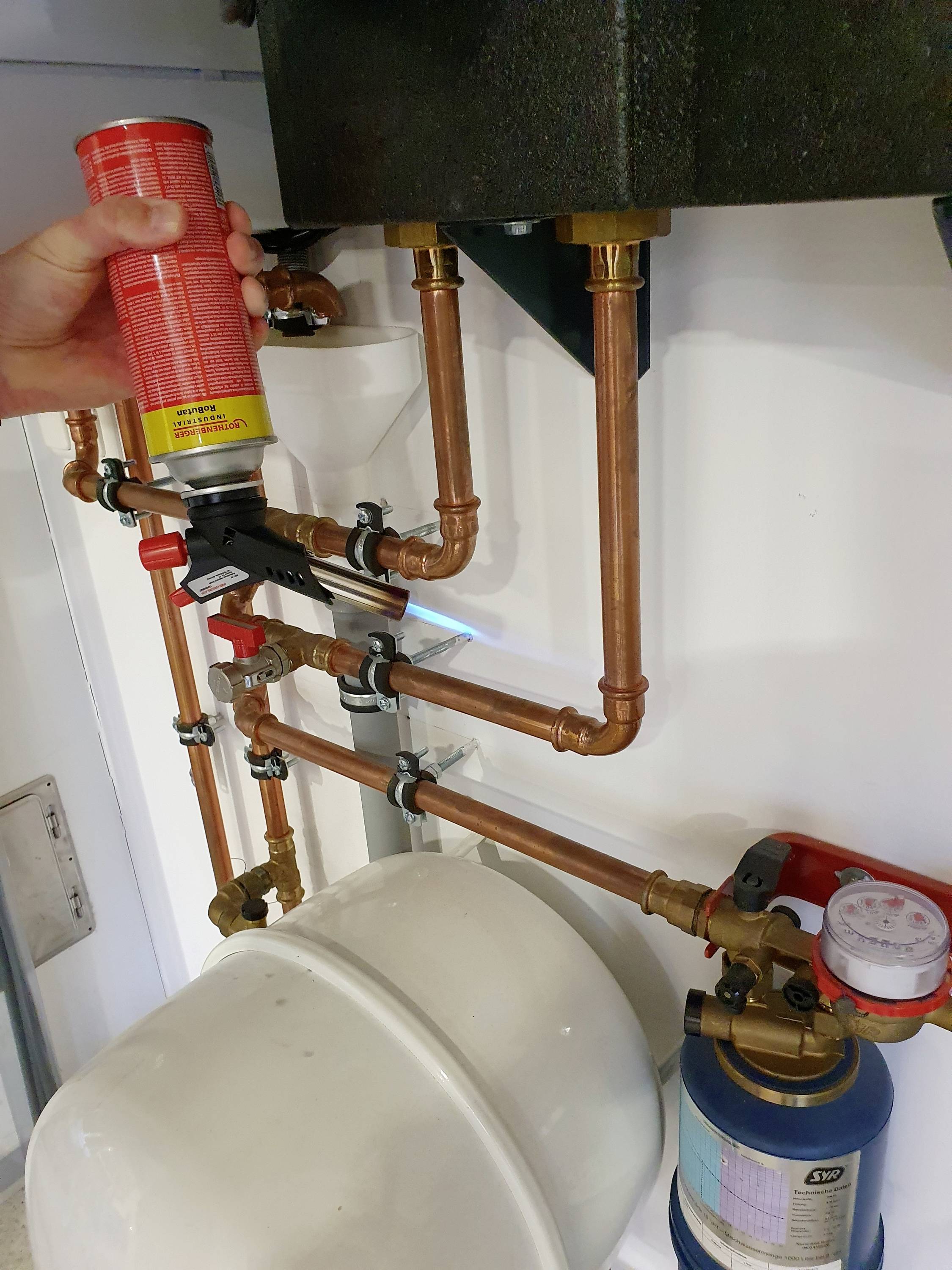 Tuyaux en cuivre et accessoires pour installations sanitaires, avec un manomètre et une personne tenant une bombe aérosol, visibles.