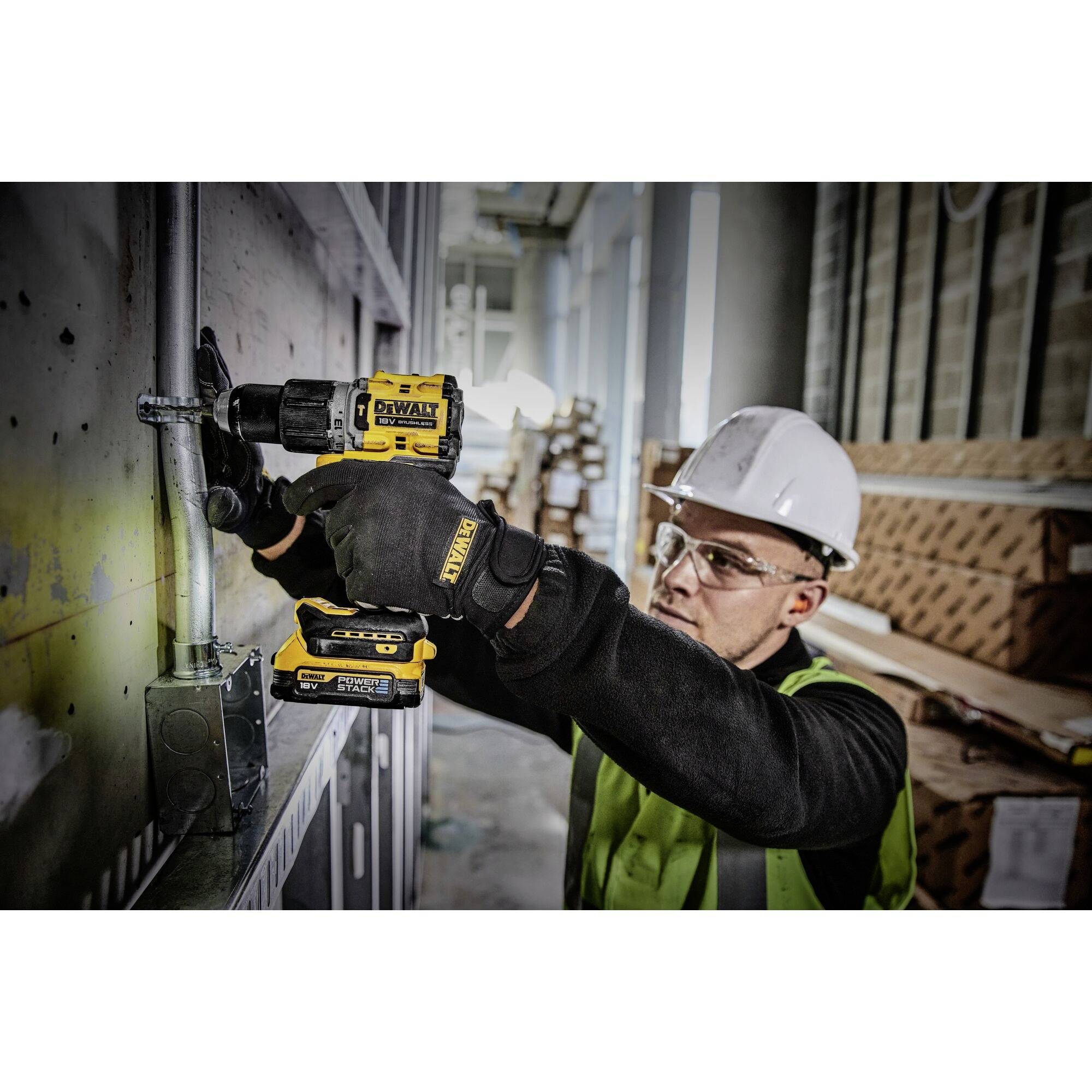 Un ouvrier portant des lunettes de protection et un casque fore un mur avec une perceuse jaune. Il porte un gilet jaune, entouré de matériel de chantier.