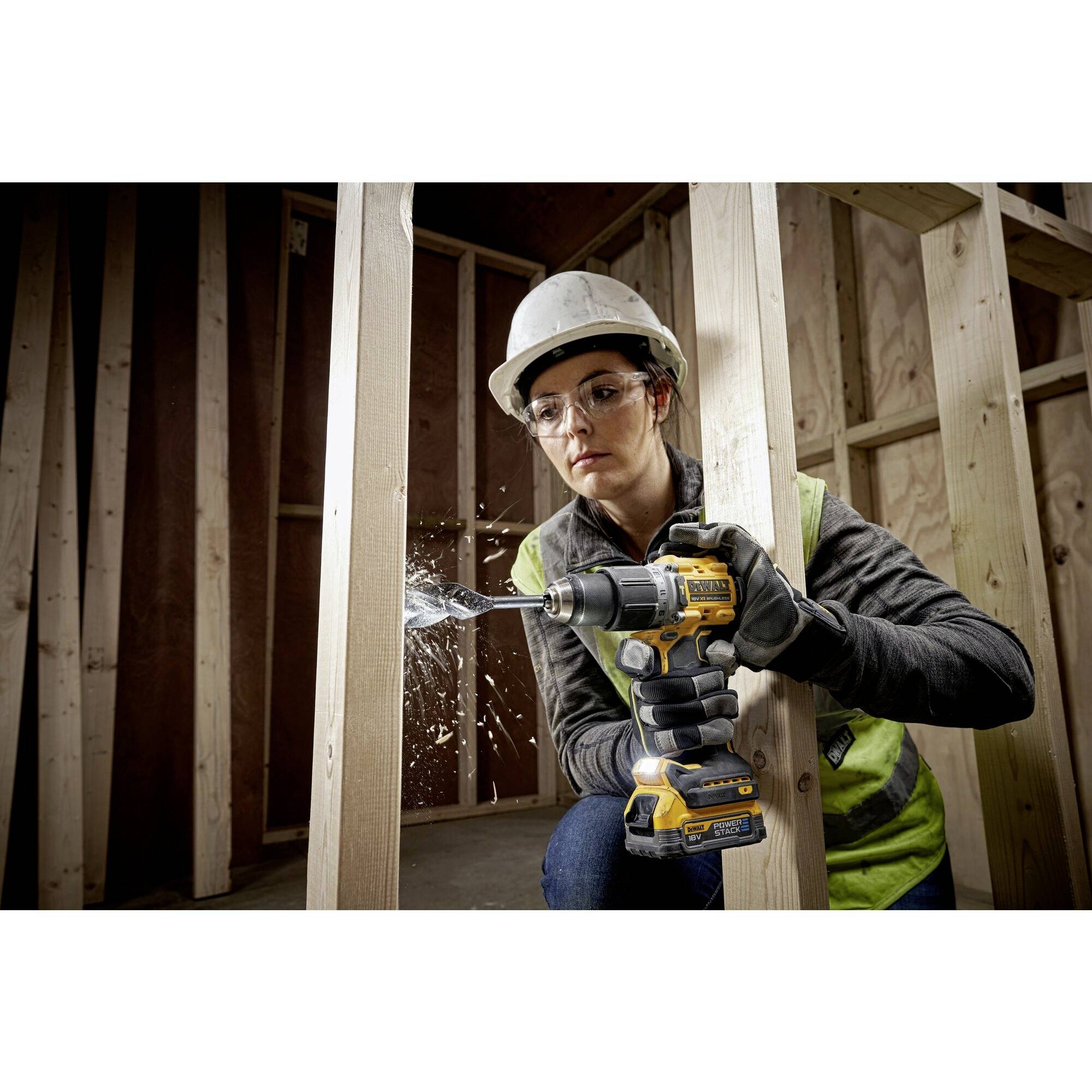 Une personne portant un casque et des lunettes de protection fore un cadre en bois à l'aide d'une perceuse électrique sur un chantier de construction.