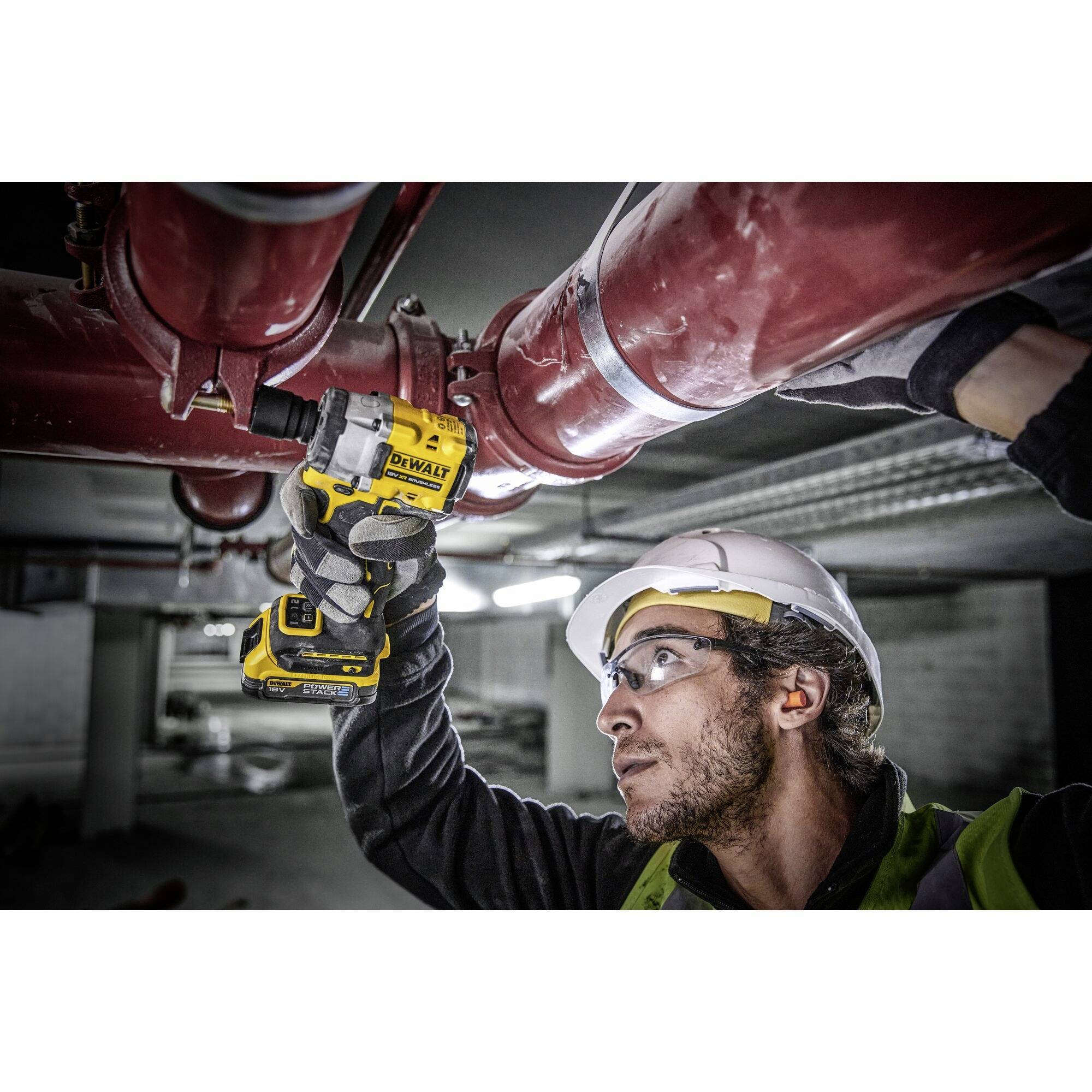 Un travailleur portant un casque de sécurité et un gilet haute visibilité utilise un tournevis électrique jaune pour travailler sur un tuyau rouge.