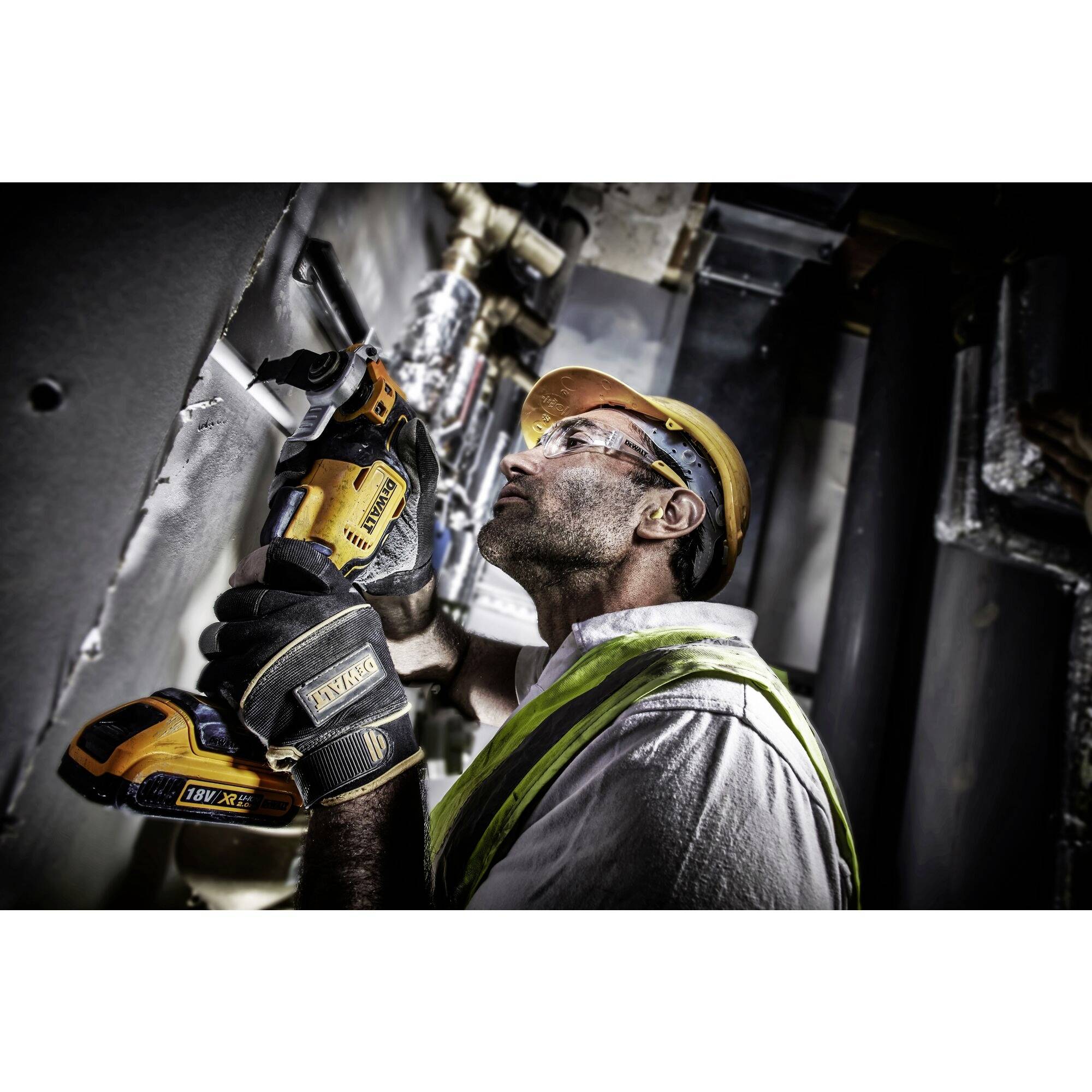 Un ouvrier portant un casque de chantier et un gilet de sécurité utilise une scie électrique pour couper un tuyau dans un espace industriel.