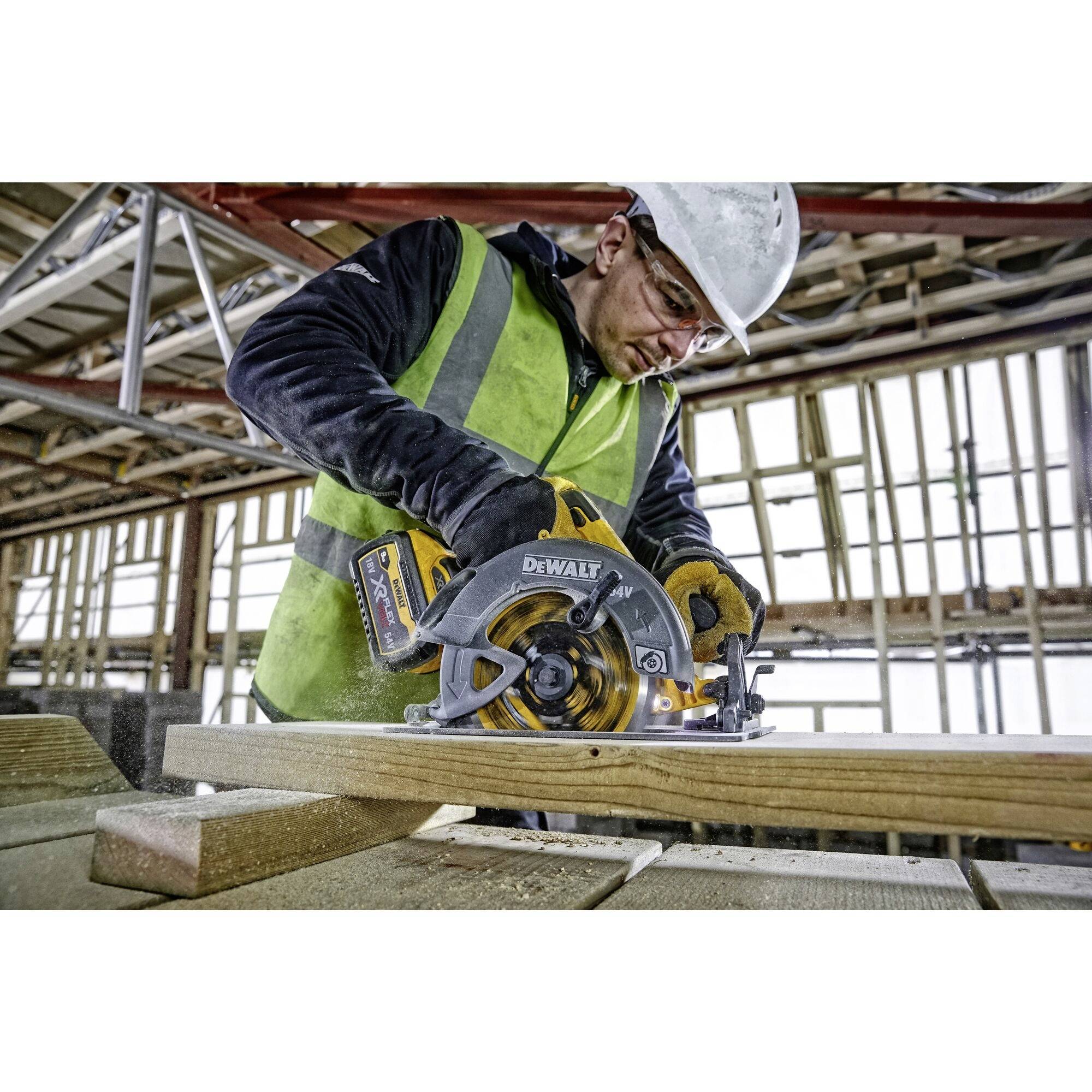 Un ouvrier portant un casque de chantier et un gilet de sécurité coupe du bois à la scie circulaire dans un bâtiment en construction. Des échafaudages sont visibles en arrière-plan.