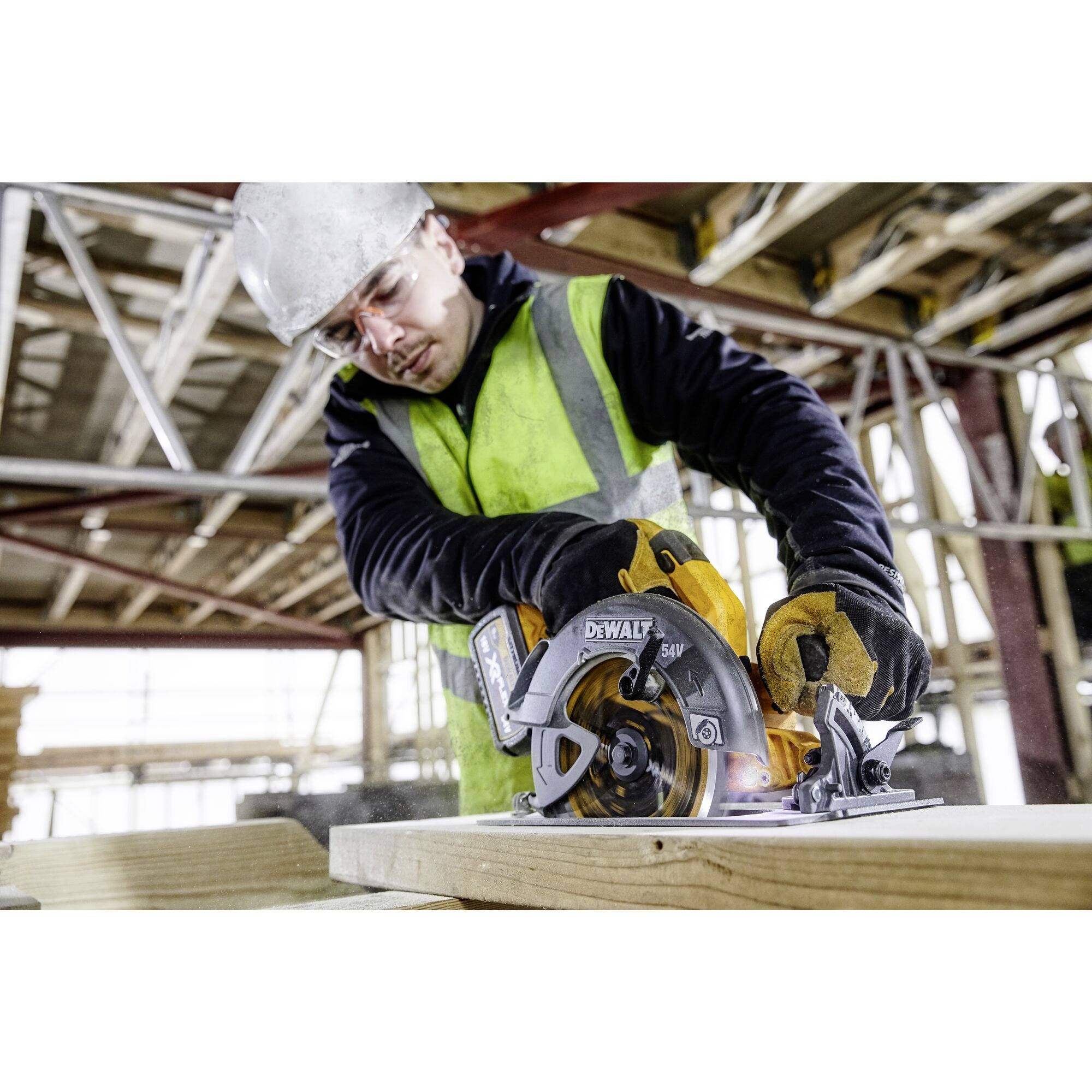 Un ouvrier portant un équipement de protection coupe du bois avec une scie circulaire sur un chantier de construction. Des échafaudages sont visibles en arrière-plan.