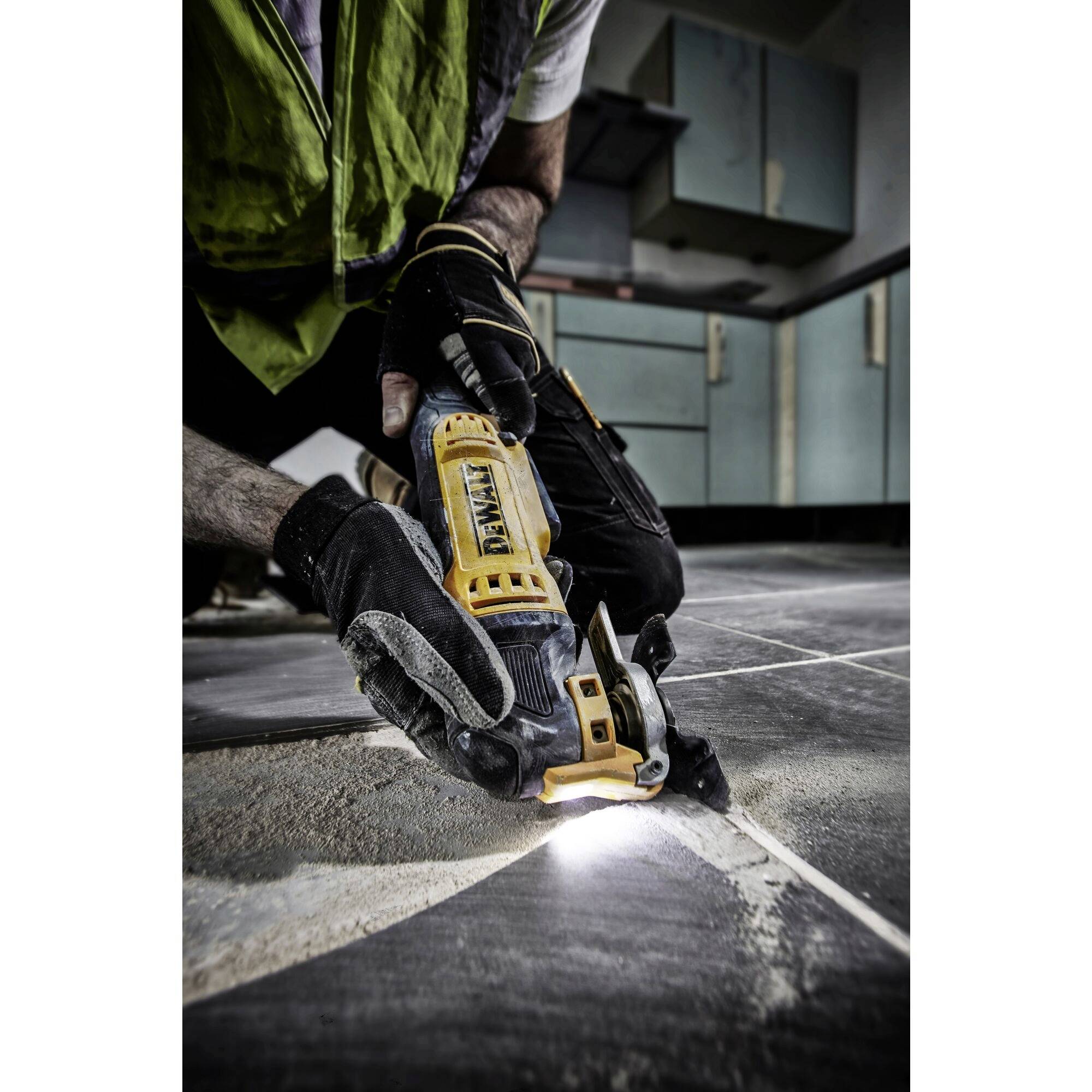 Un ouvrier portant des gants de protection utilise un outil électrique pour couper des carreaux dans une cuisine. De la poussière et de la lumière sont visibles.