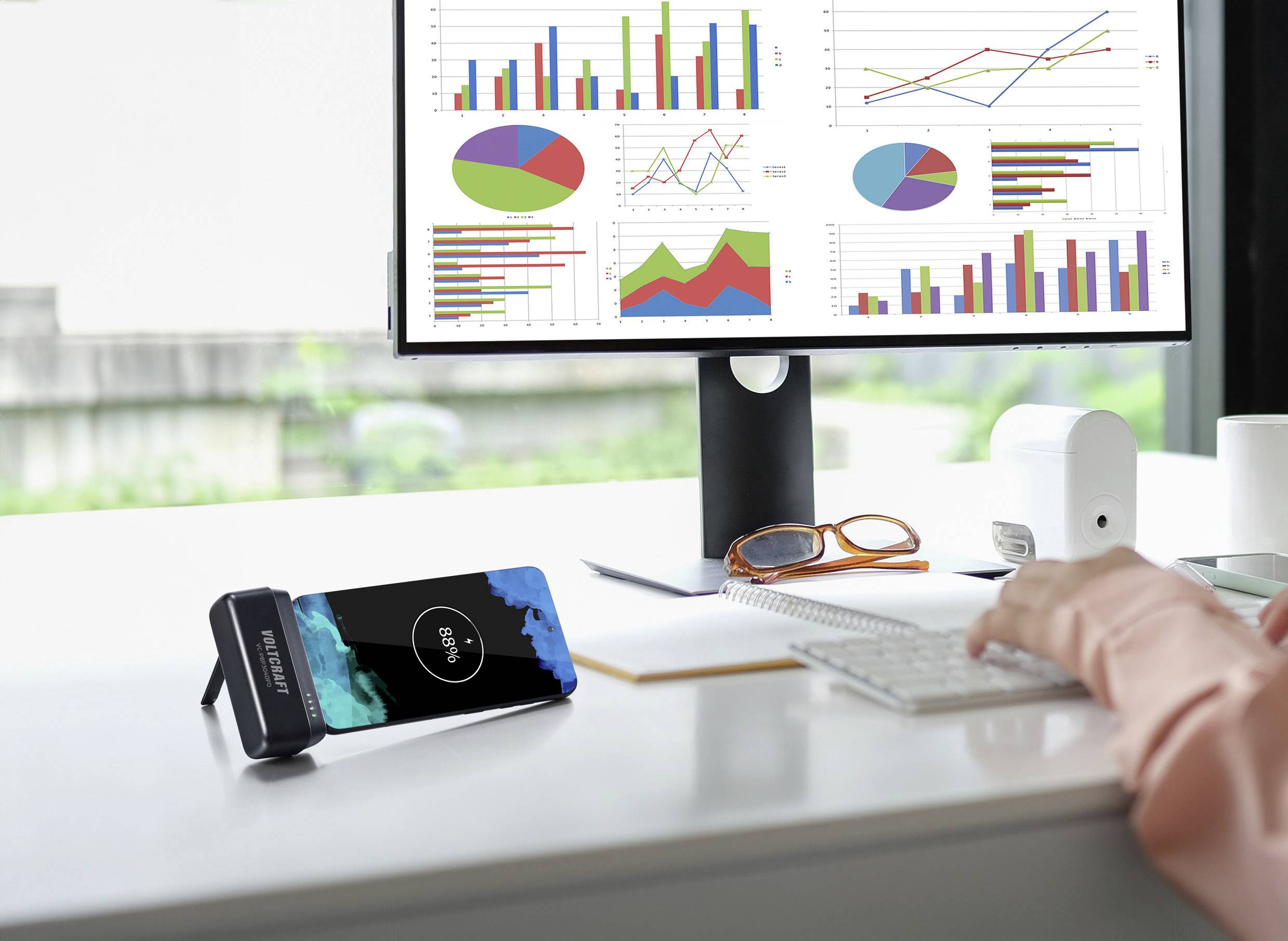 Une personne travaille à un bureau avec des graphiques sur un écran d'ordinateur. Un smartphone est en train de charger sans fil sur un support.