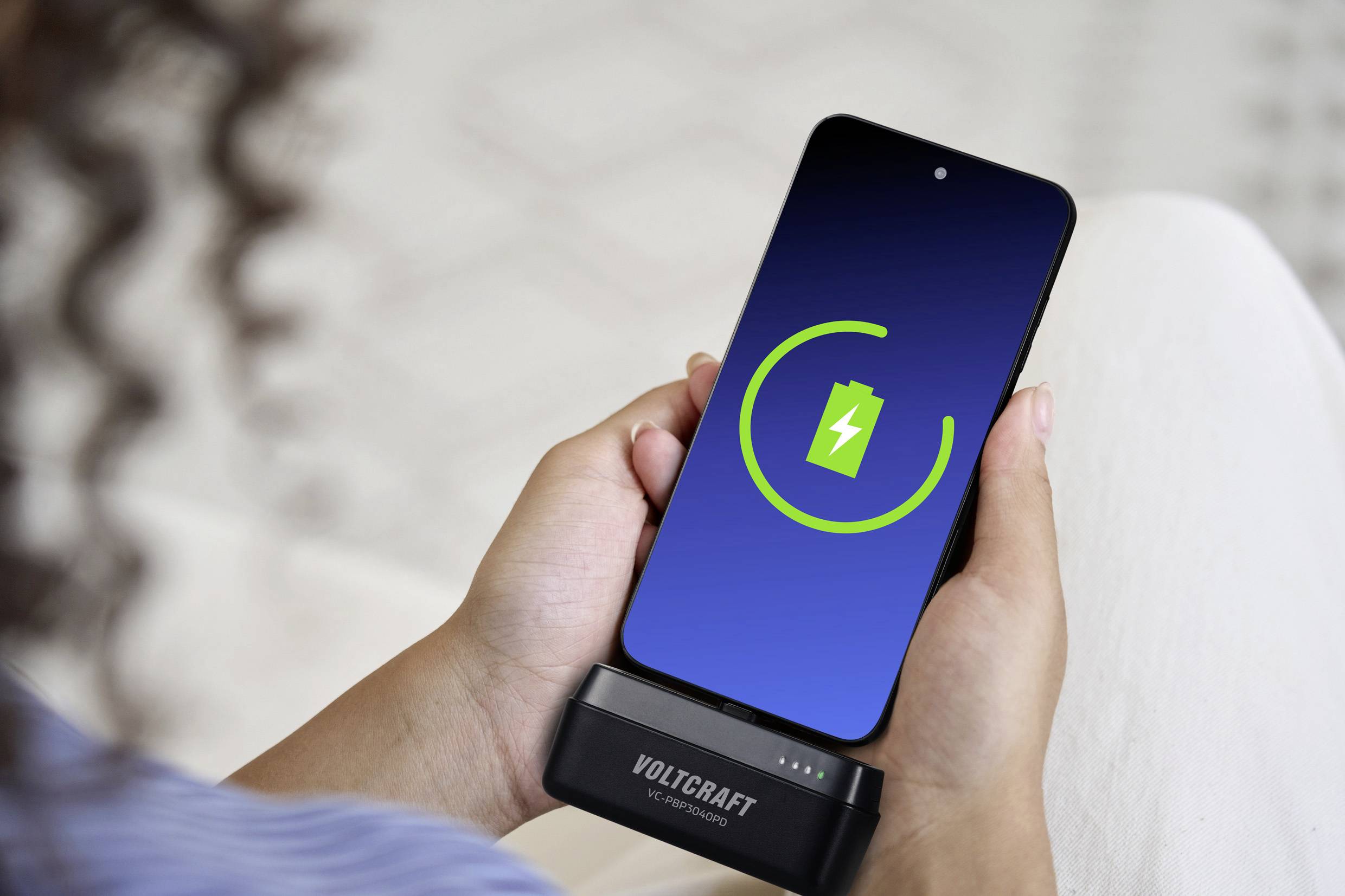 Une personne tient un smartphone avec un indicateur de charge à l'écran. L'appareil est en cours de chargement avec un chargeur VOLTCRAFT.