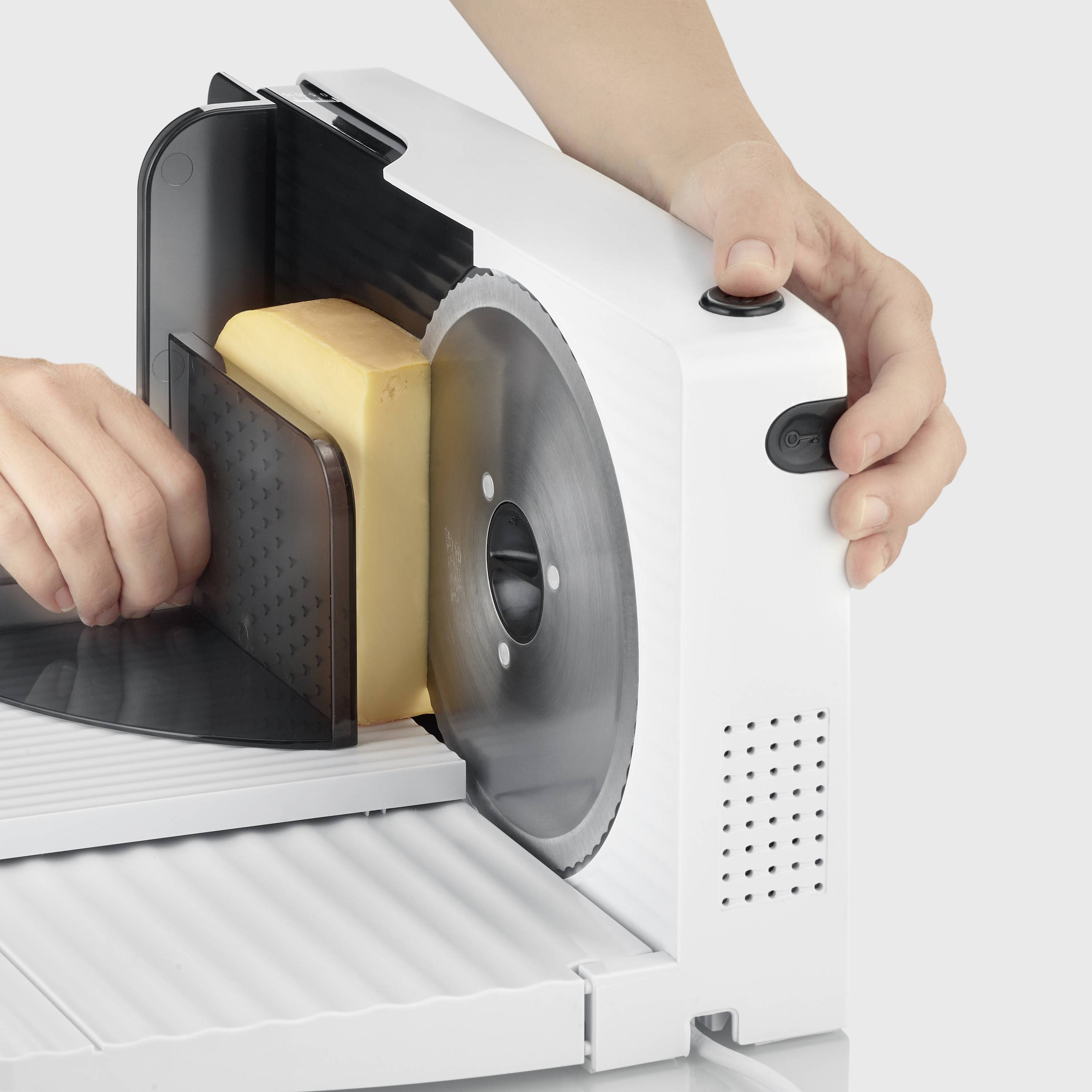 Une main coupe un grand morceau de fromage en tranches à l'aide d'une trancheuse électrique. À côté de la tranche se trouve un volant à main.