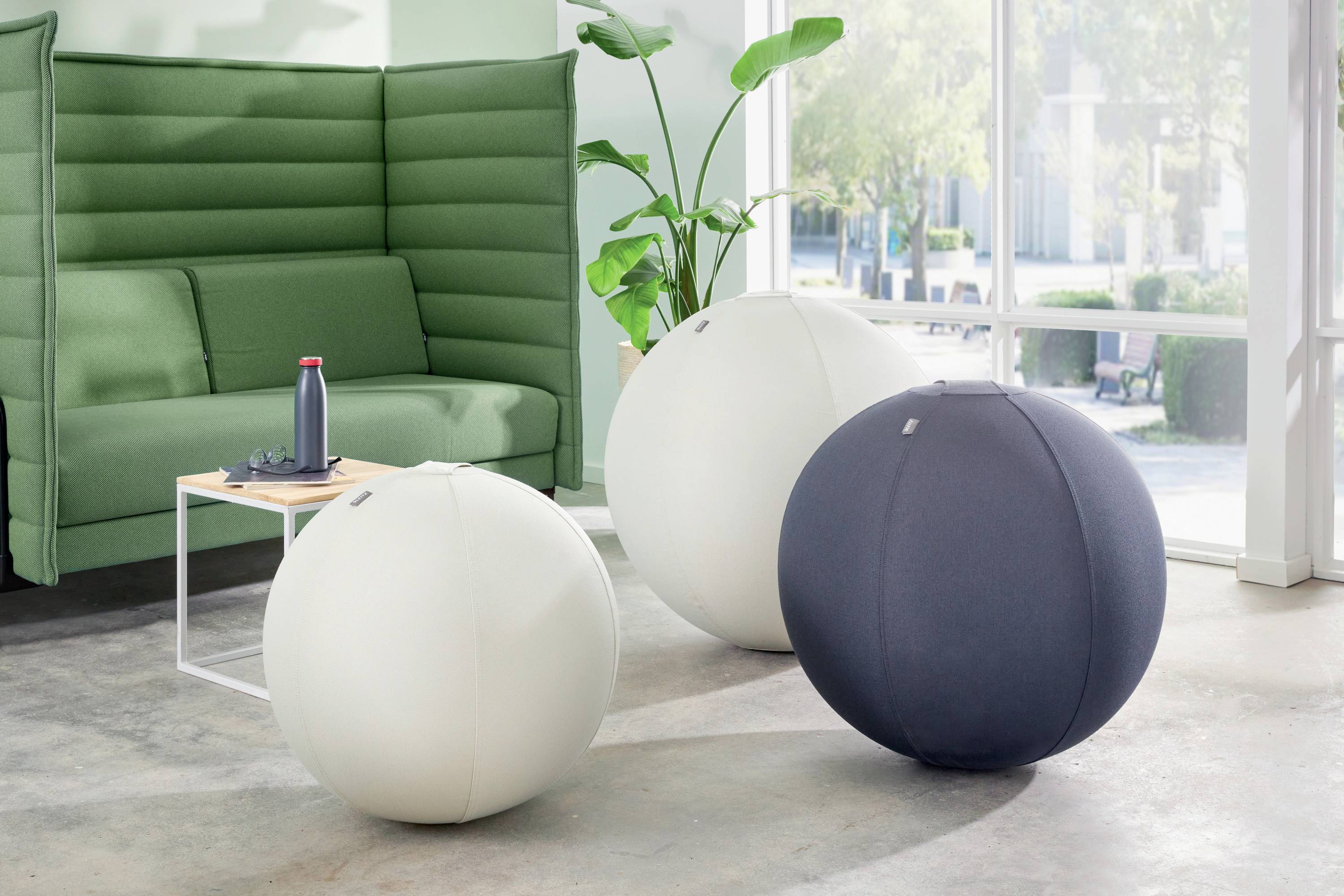 Trois grands ballons de gymnastique, deux blancs et un bleu foncé, devant un canapé vert et une plante dans une pièce claire.