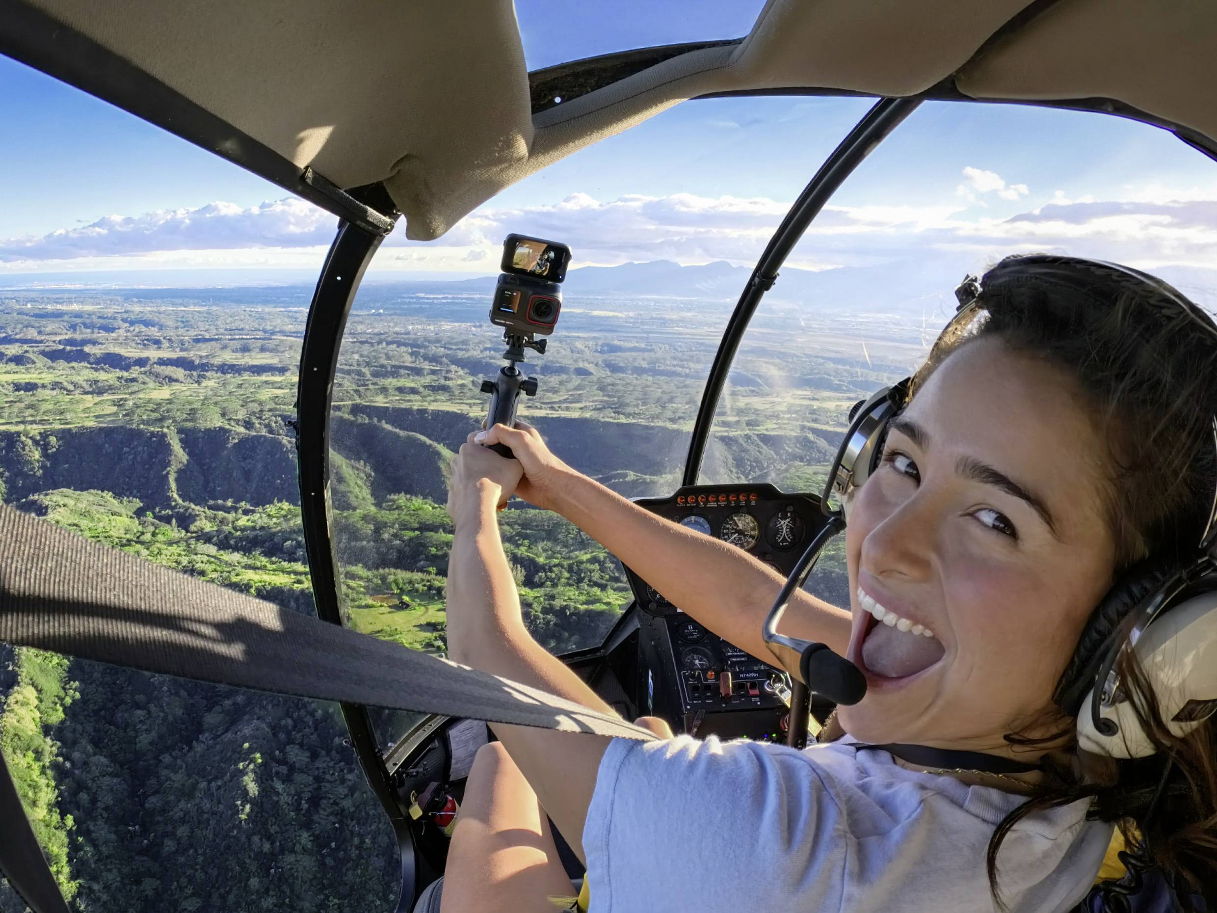 Une personne souriante est assise dans un hélicoptère, tenant une caméra GoPro et survolant un paysage verdoyant avec des montagnes en arrière-plan.