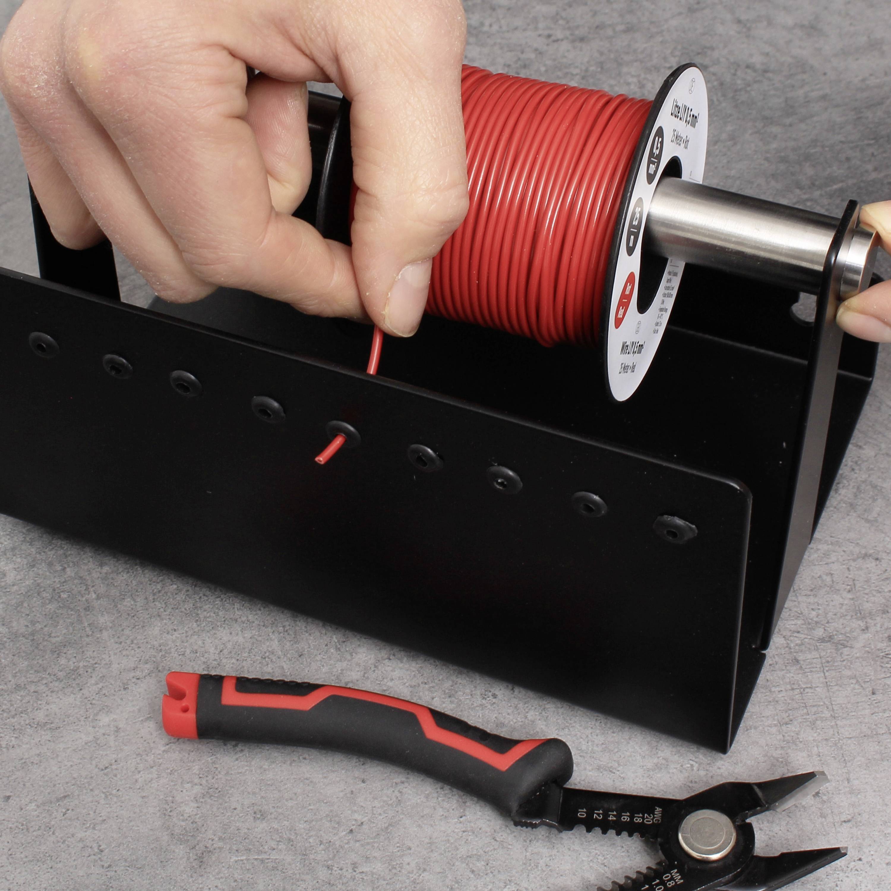 Une main tient un rouleau de câble rouge monté sur un support ; à côté, un coupe-câble avec une poignée rouge est posé sur une surface grise.