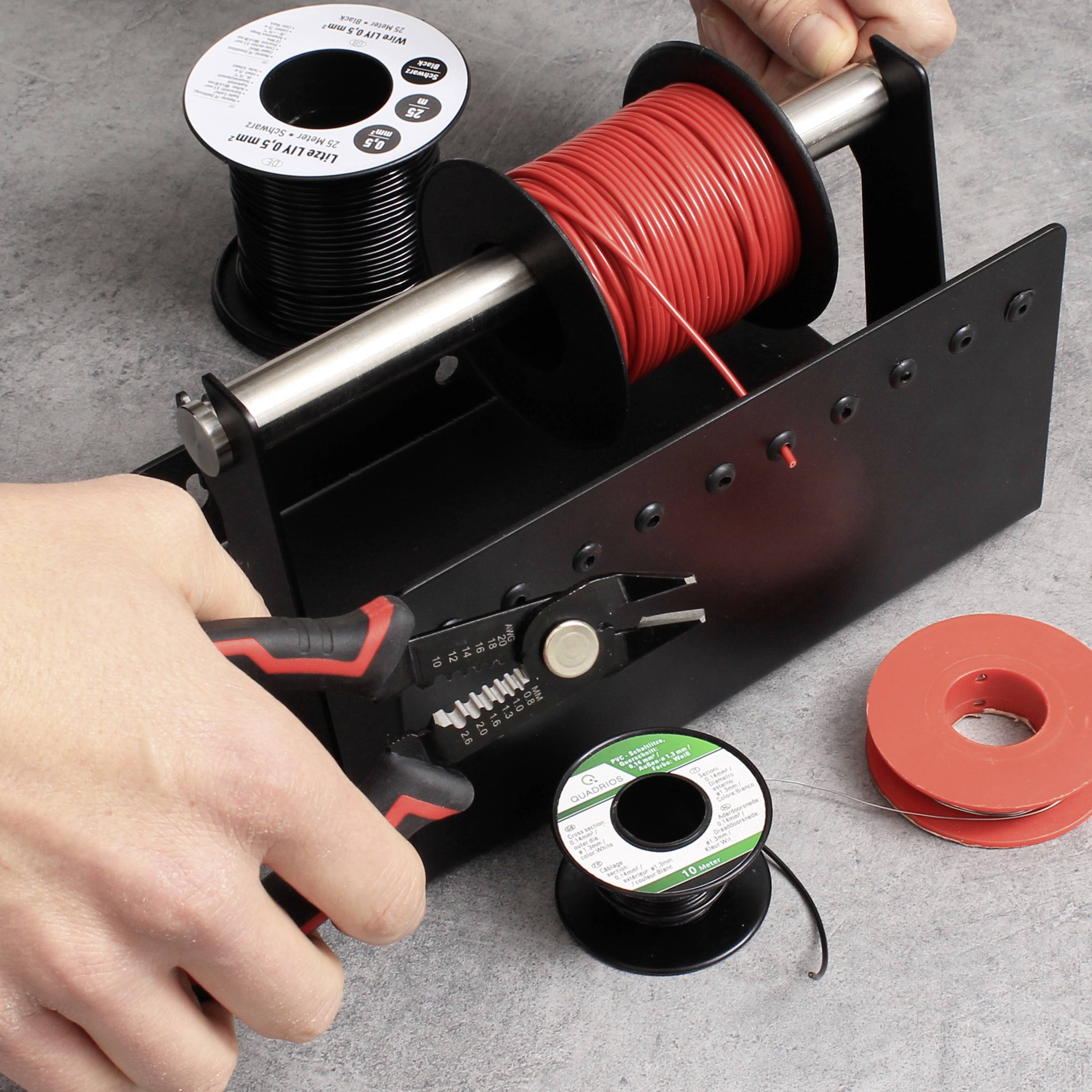 Une main tient une pince et coupe un câble rouge d'une bobine sur un support, avec d'autres rouleaux de ruban isolant à côté. Poste de travail d'un électricien.