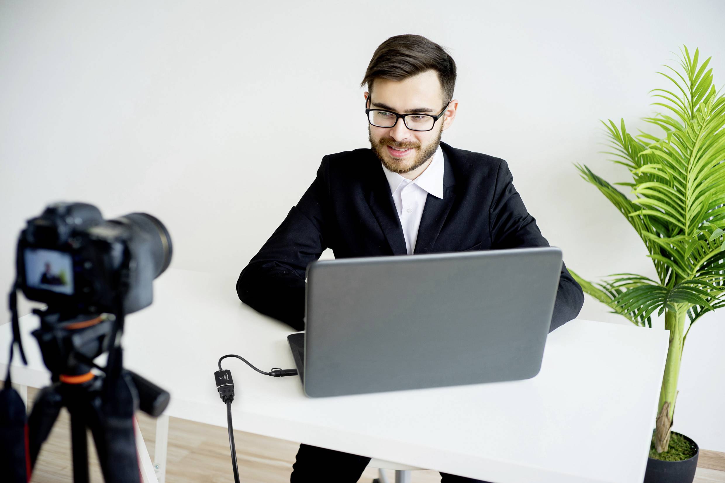 Un homme en costume noir est assis à une table devant un ordinateur portable, regardant dans une caméra. Une plante se trouve à sa droite.