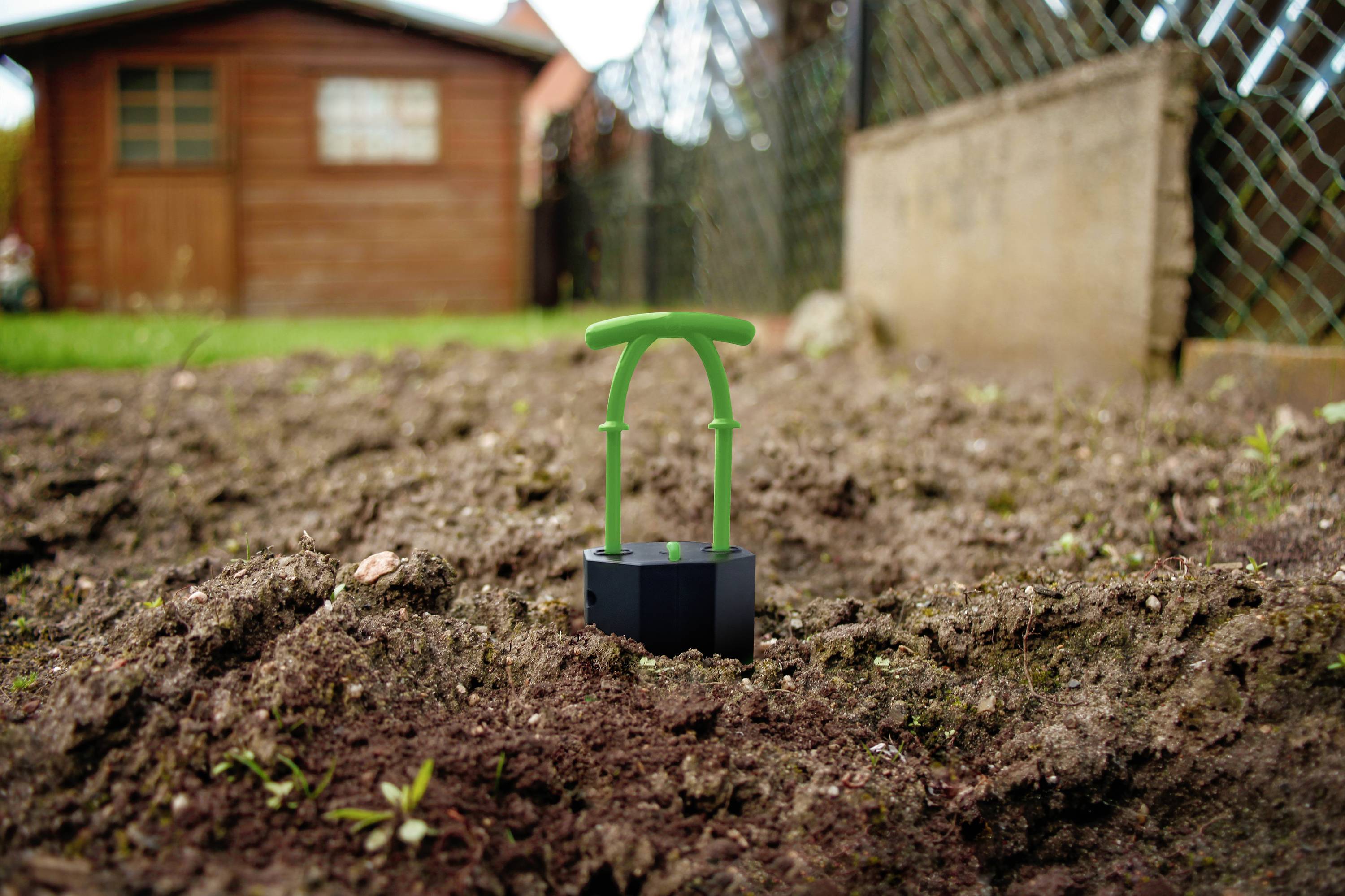 Un sol de jardin avec un petit dispositif d'aide à la plantation vert planté dans la terre. À l'arrière-plan, un abri de jardin en bois.