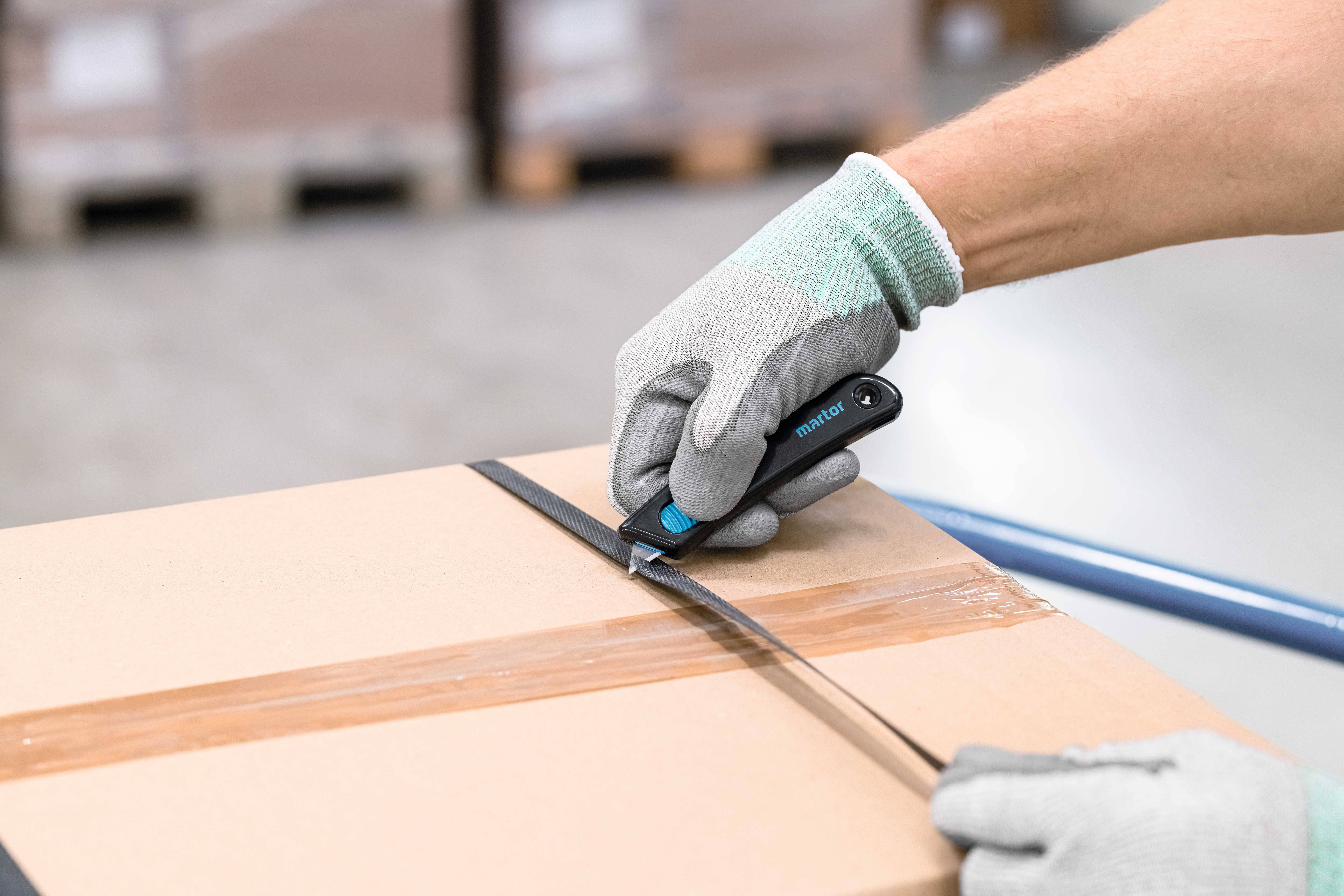 Une personne portant des gants gris coupe du ruban adhésif sur un carton à l'aide d'un couteau utilitaire. Des palettes floues sont visibles en arrière-plan.