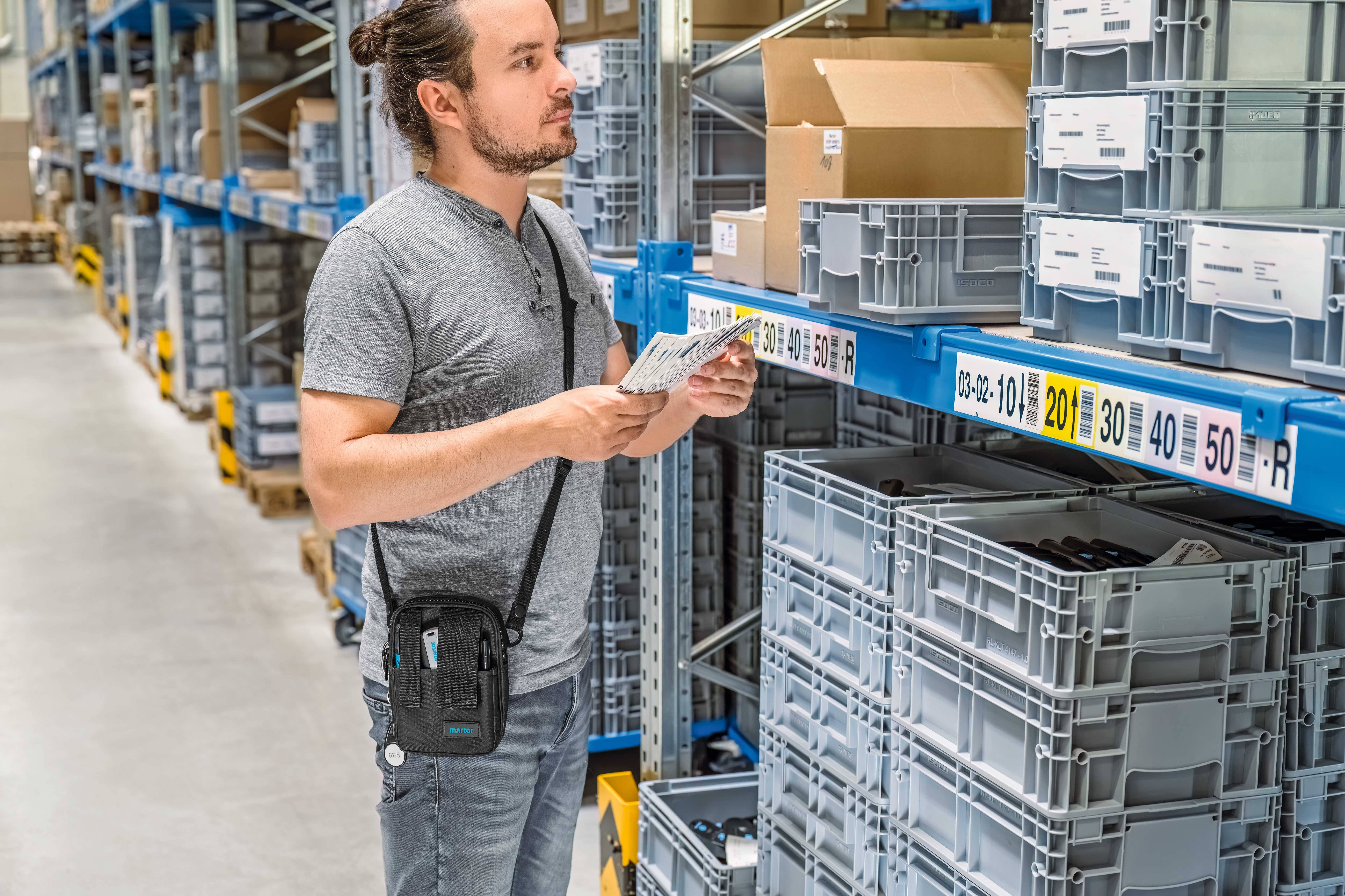 Un homme scanne des produits dans un entrepôt. Il porte un t-shirt gris et un sac. Les étagères sont remplies de caisses numérotées.