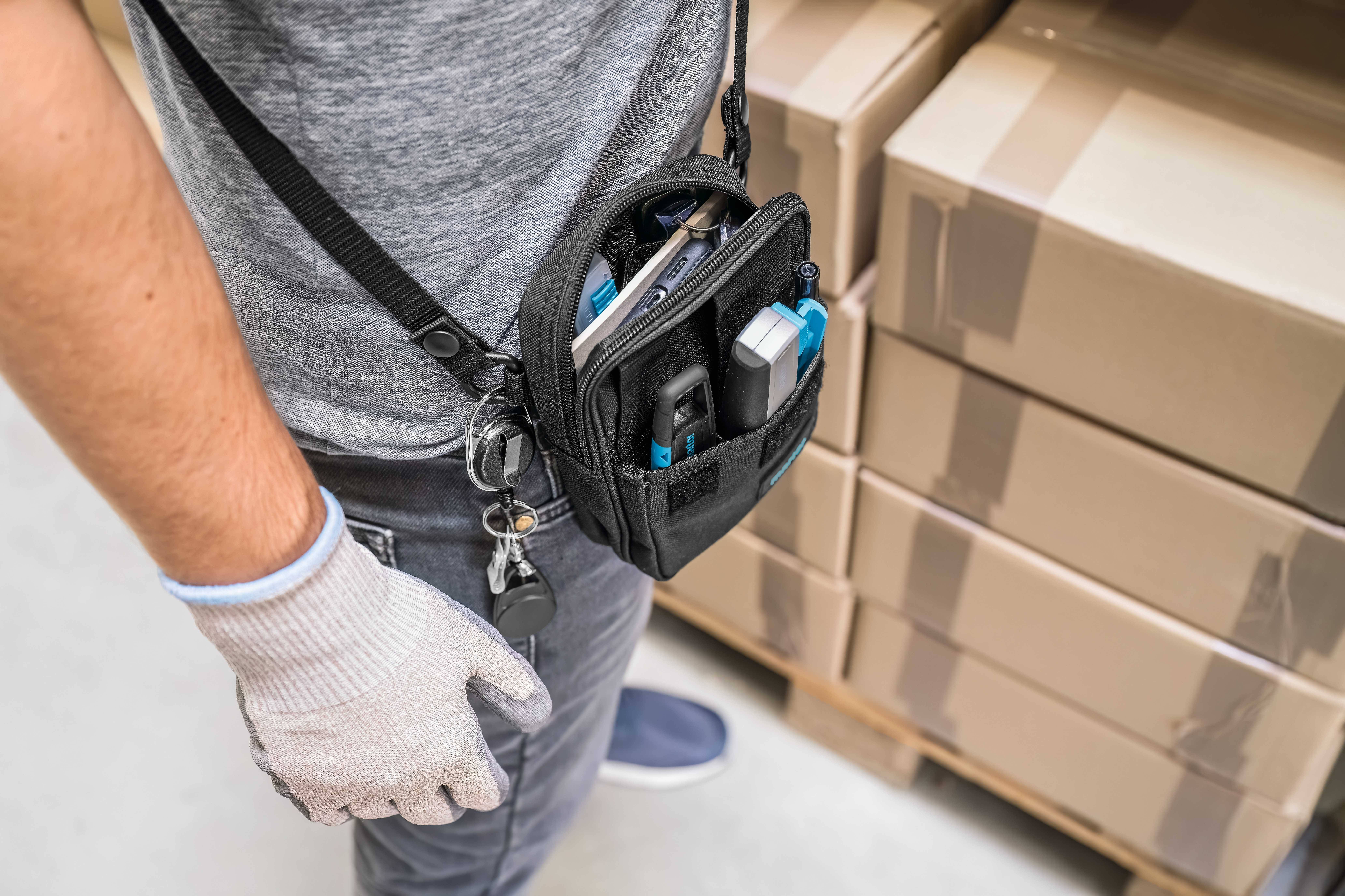 Une personne porte un sac à outils et accessoires, debout devant des cartons empilés dans un environnement d'entrepôt.