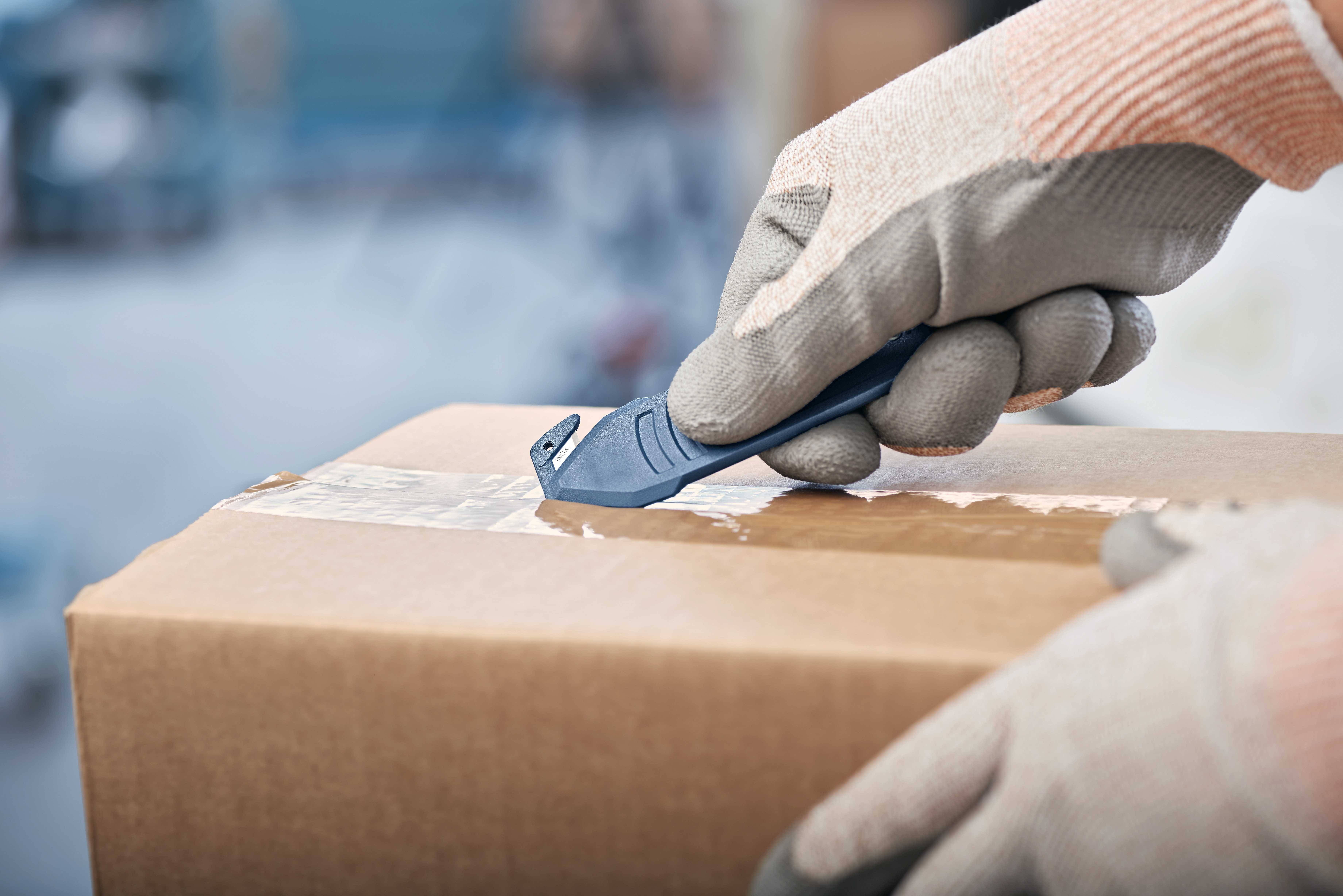Une personne portant des gants de travail ouvre un colis scellé à l'aide d'un couteau. Le focus est mis sur le mouvement précis de coupe.