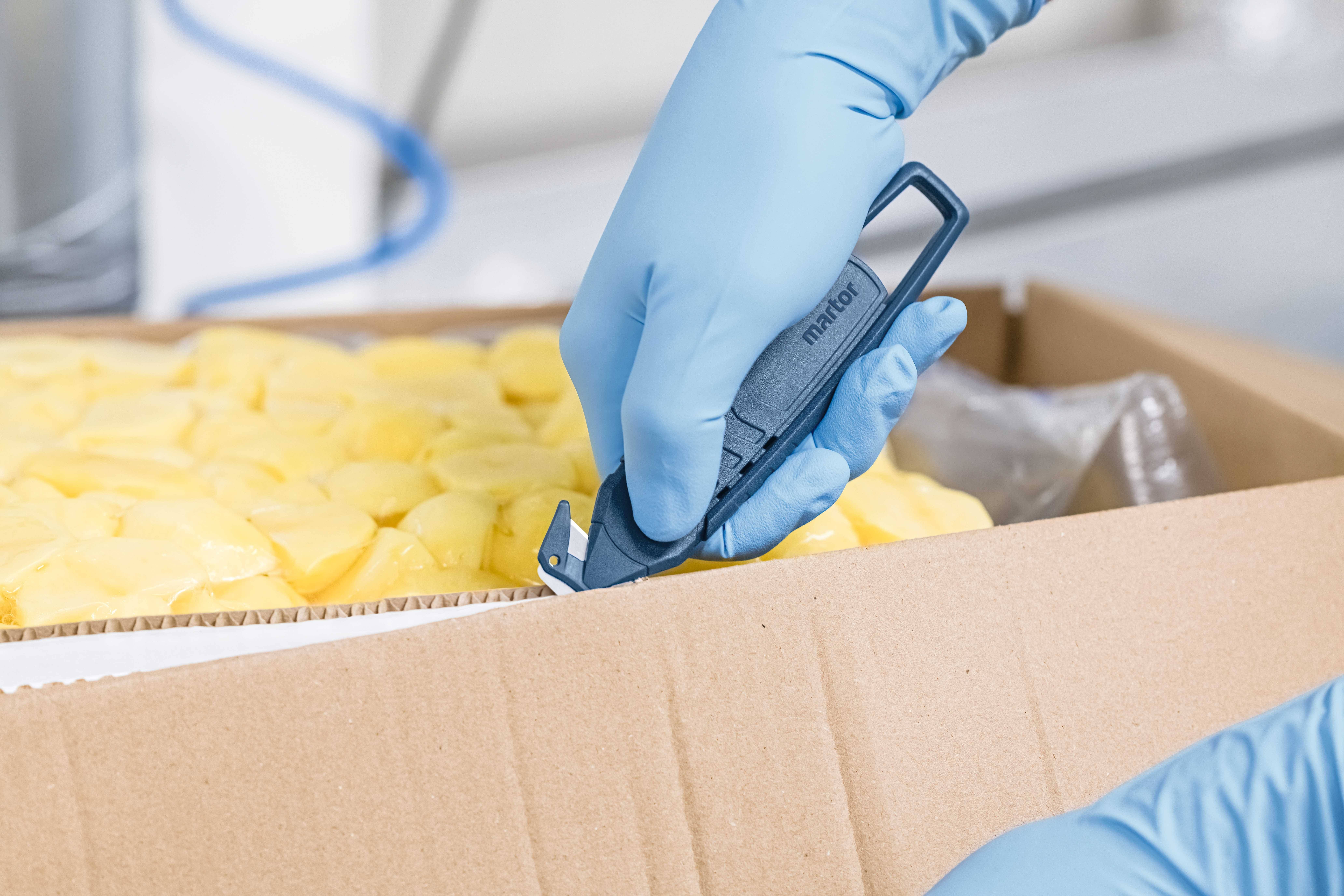 Une main gantée de bleu ouvre un carton rempli d'aliments jaunes à l'aide d'un outil de coupe.