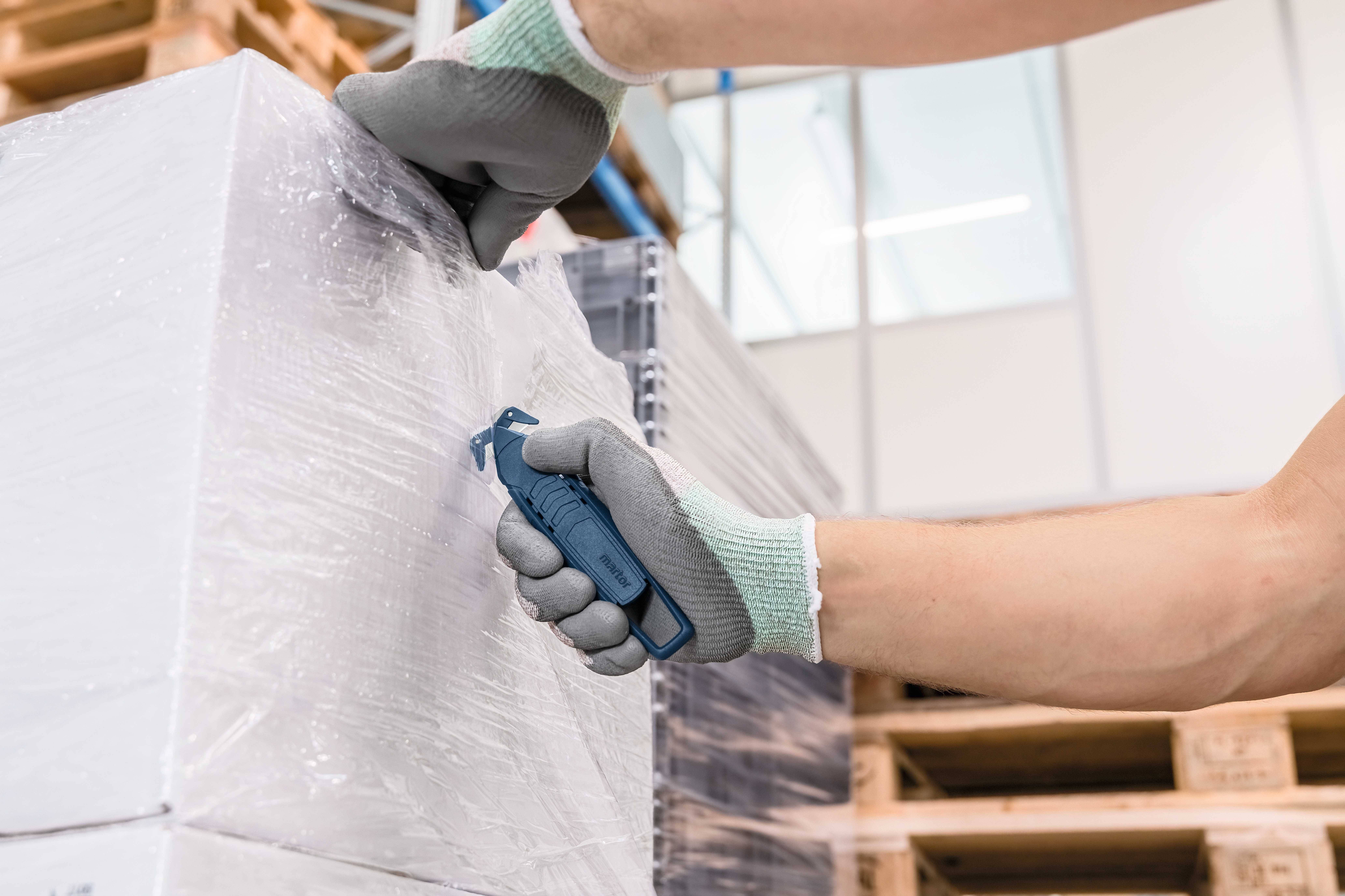 Une personne coupe le film plastique d'une palette avec un couteau de sécurité. Des gants protègent ses mains.