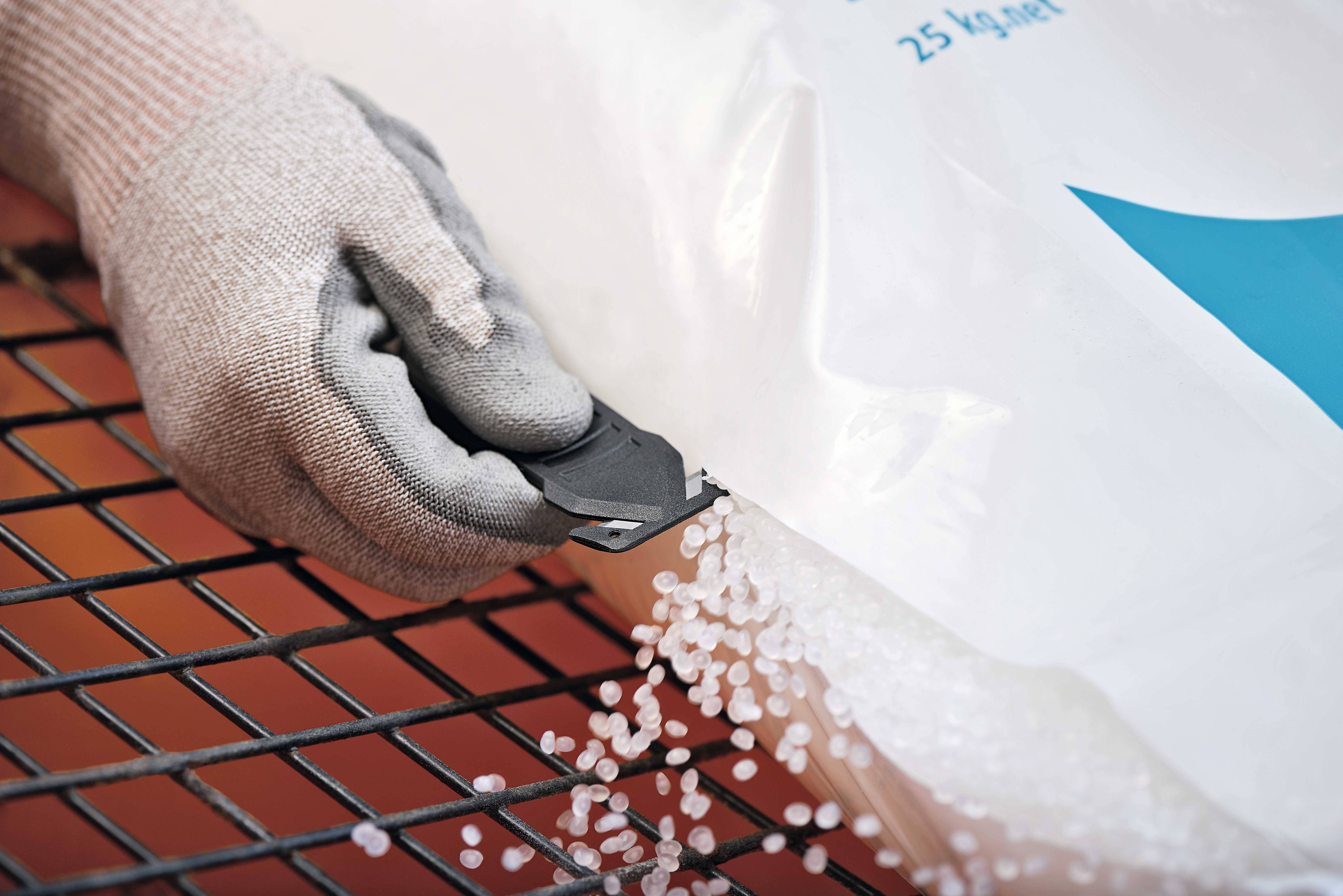 Une main gantée découpe un sac en plastique avec un couteau, et des granulés de plastique s'écoulent sur une table grillagée.