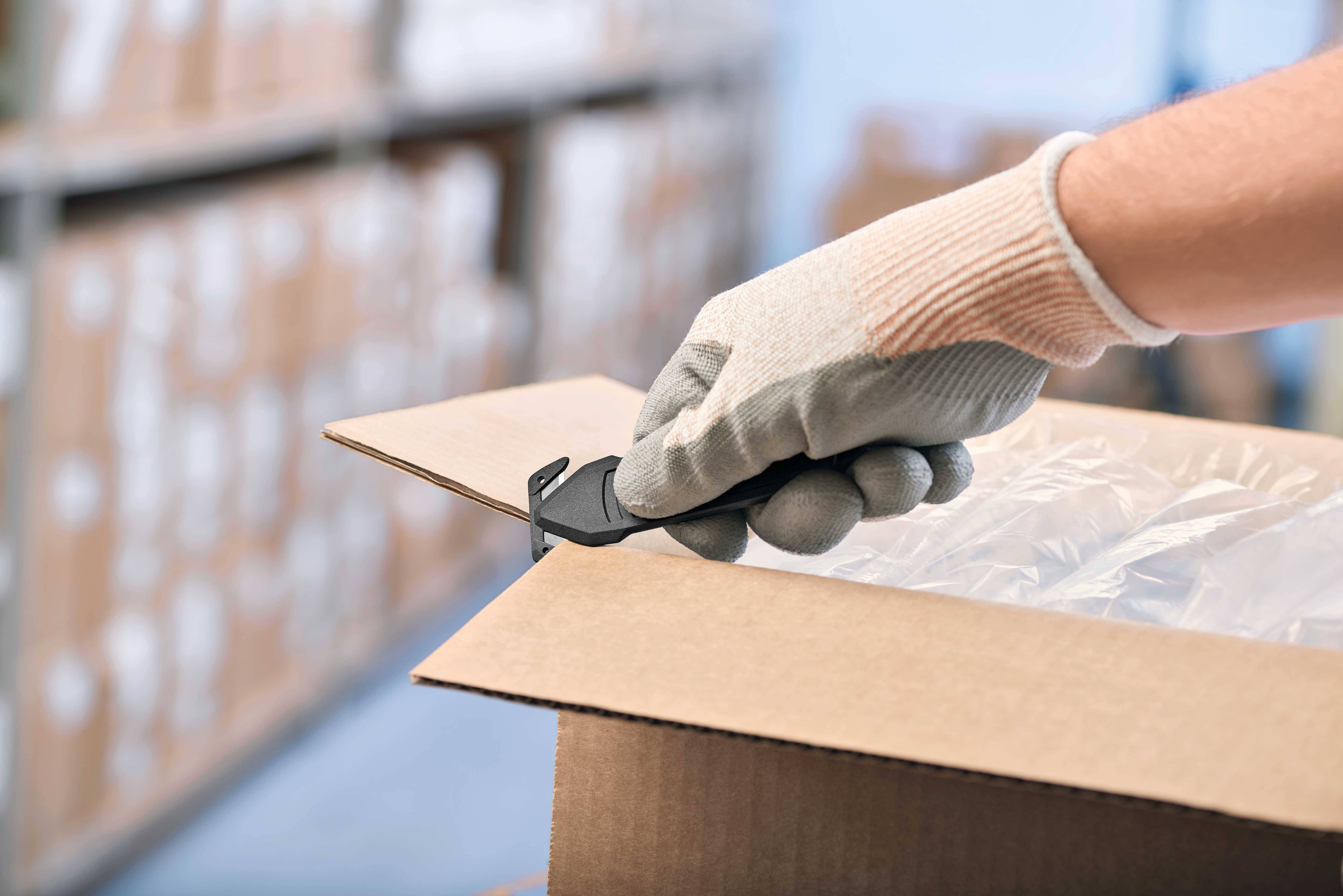Une personne portant des gants ouvre un carton à l'aide d'un couteau de sécurité. L'arrière-plan montre des étagères de stockage floues avec d'autres cartons.