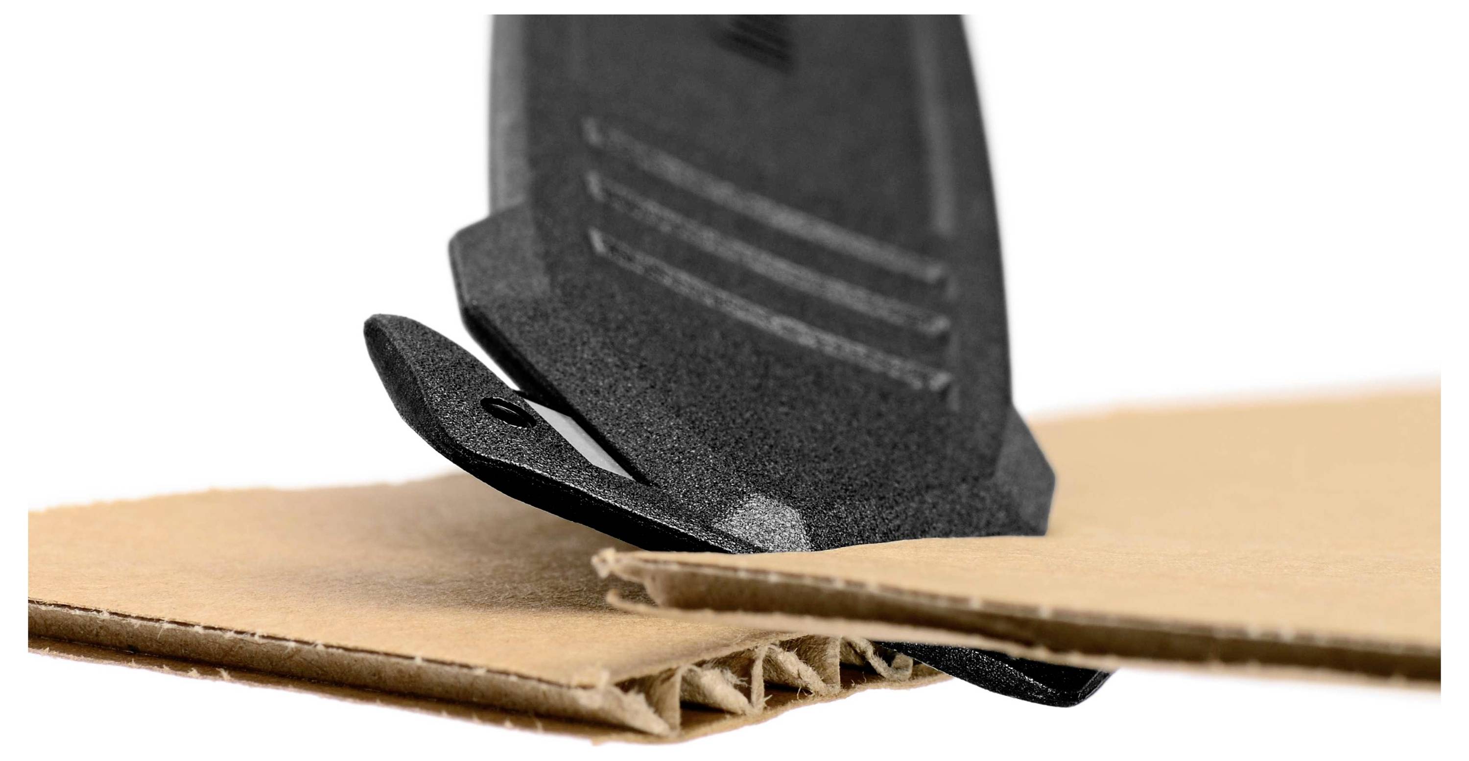 Un outil de coupe noir traverse du carton ondulé. L'outil dispose d'une lame tranchante et est positionné en biais par rapport au carton.