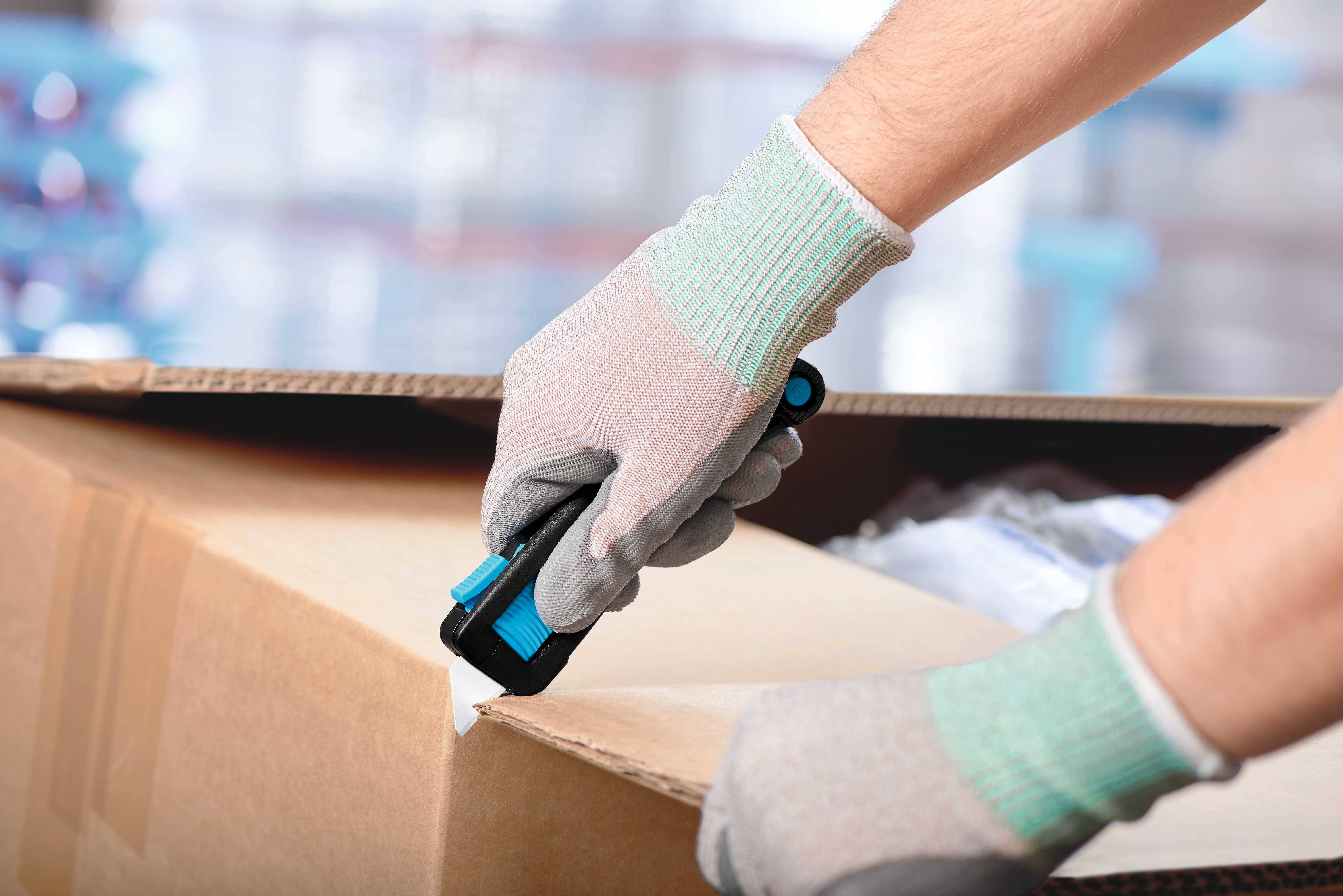Une personne ouvre un carton avec un couteau. Elle porte des gants gris. Des étagères d'entrepôt floues sont visibles en arrière-plan.