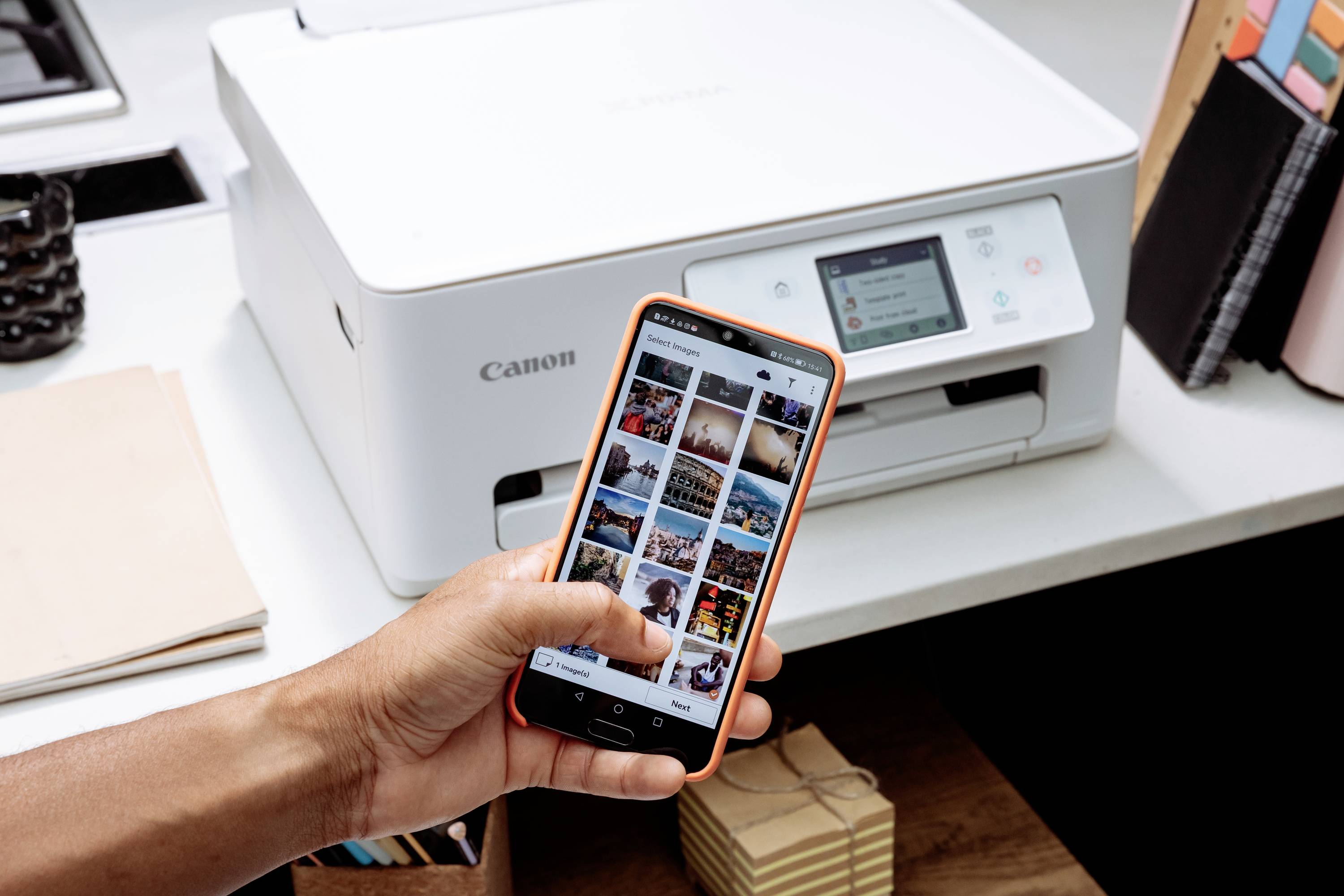 Une personne tient un smartphone devant une imprimante. Des photos sont visibles sur l'écran du téléphone. L'imprimante est posée sur une table.