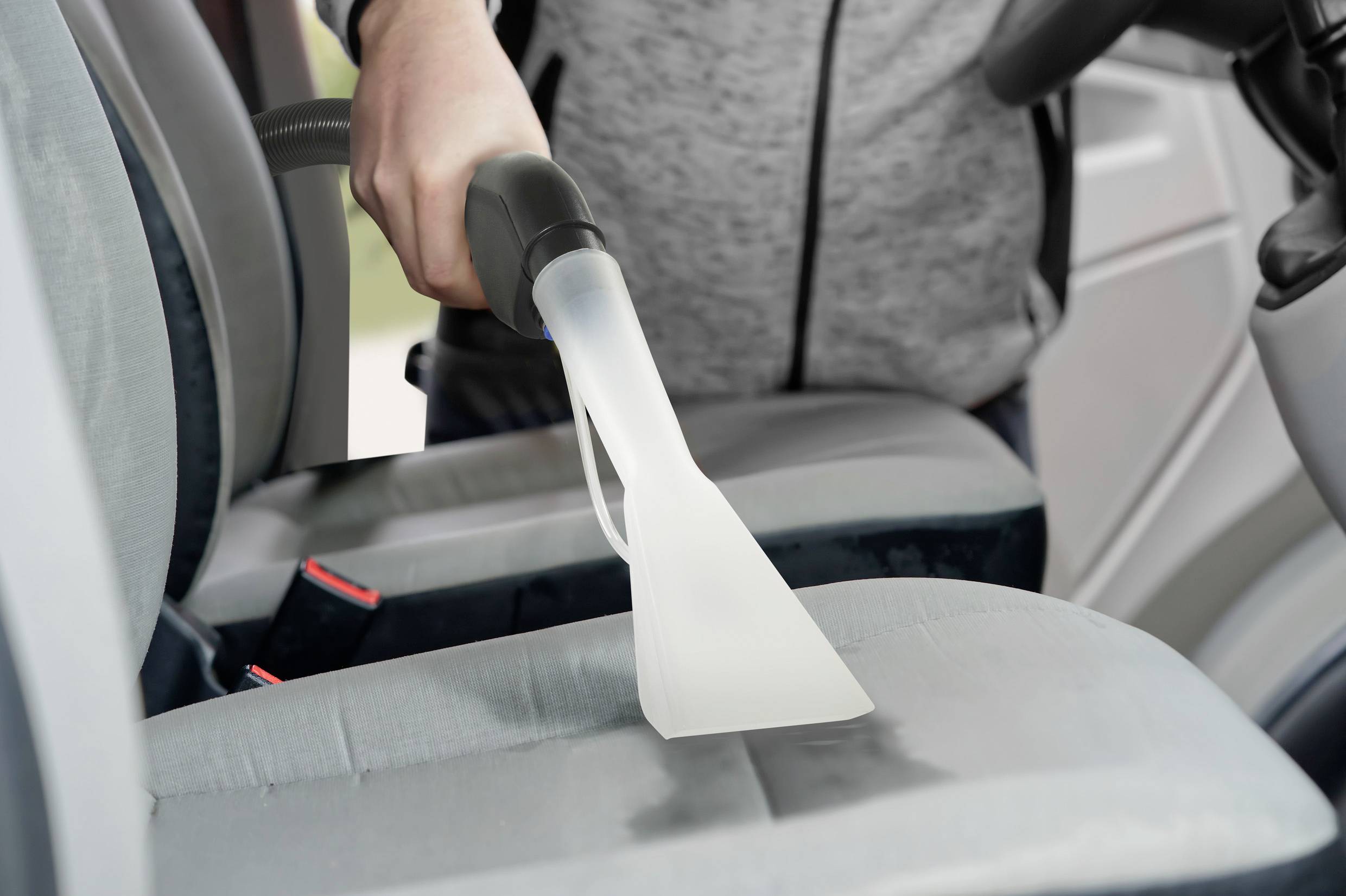Une personne nettoie le siège de voiture avec un aspirateur à eau. Le siège gris est débarrassé des taches. La scène transmet un sentiment de propreté.