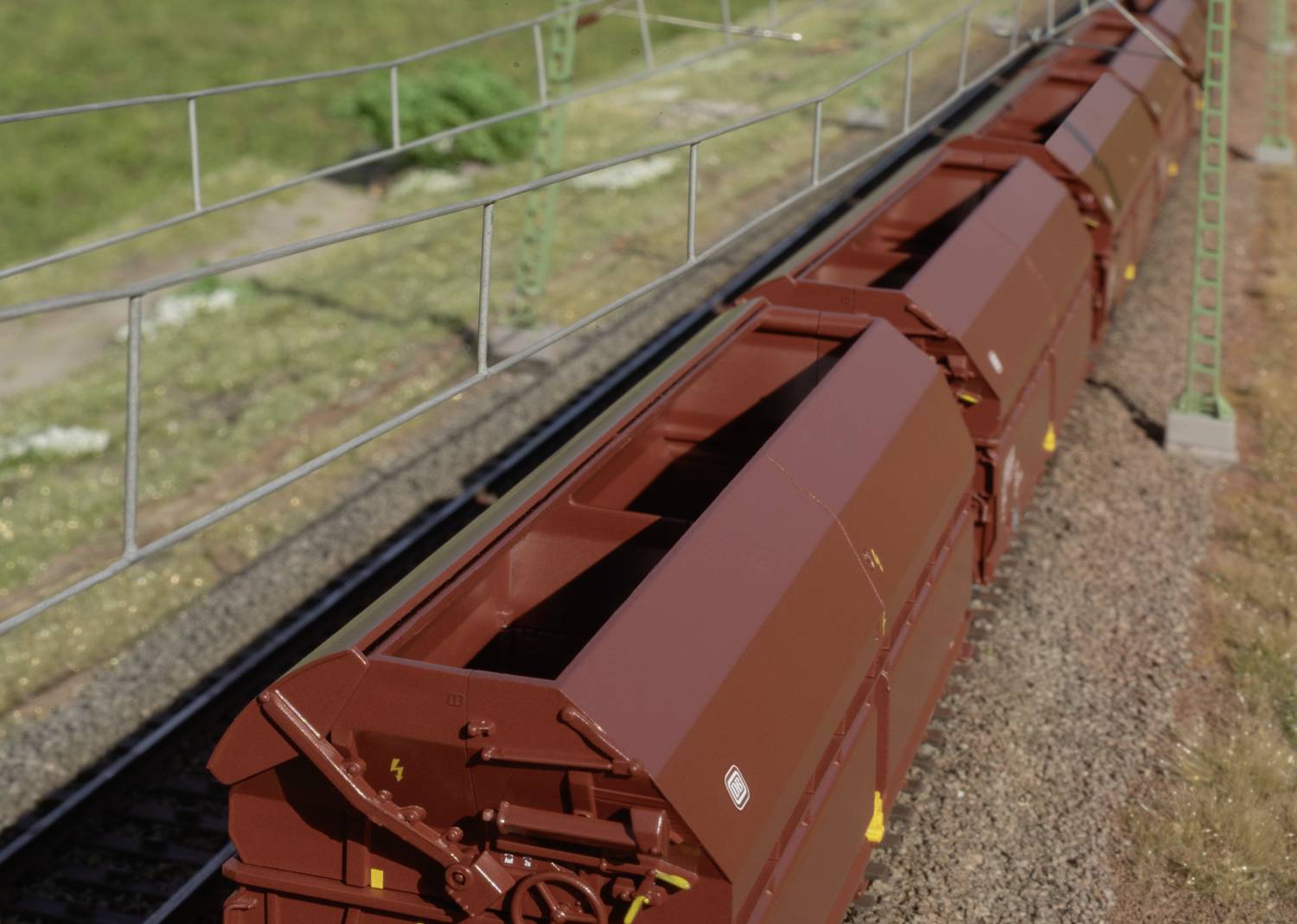 Train de marchandises avec des wagons bruns roulant sur une voie ferrée. L'environnement est composé d'herbe et d'une clôture électrique. Photographié en plongée oblique.
