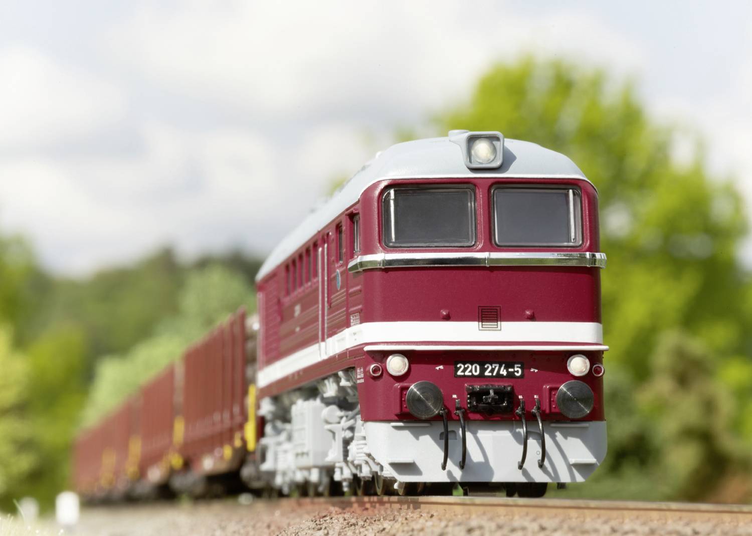 Un train de marchandises rouge circule sur un rail dans un paysage verdoyant. Au premier plan, on peut voir la locomotive portant le numéro '220 274-5'.