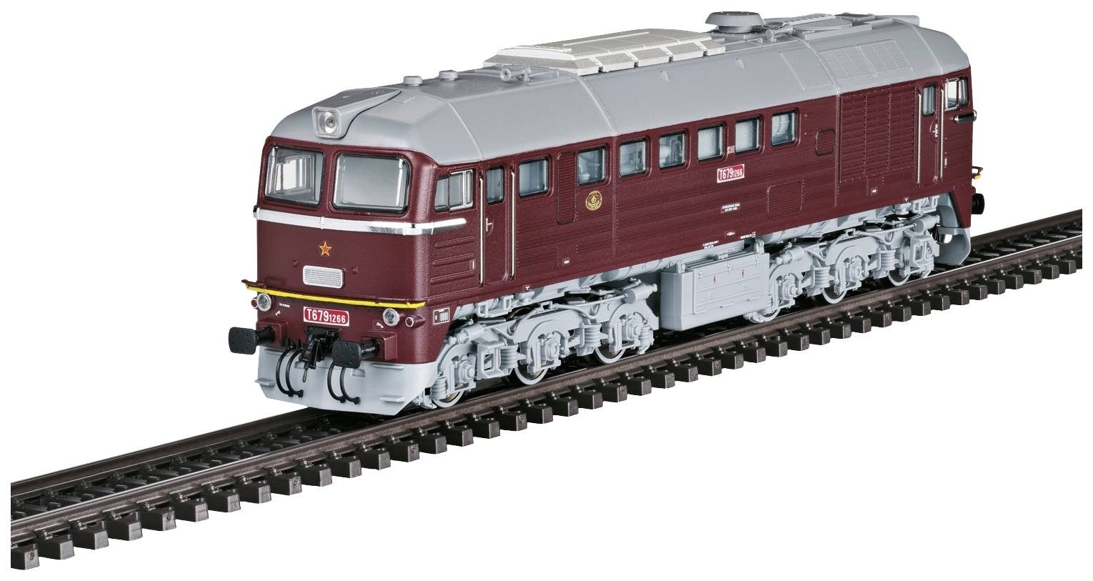 Un train miniature rouge foncé avec un toit gris roule sur des rails. Le train a une partie avant rectangulaire et des roues argentées.