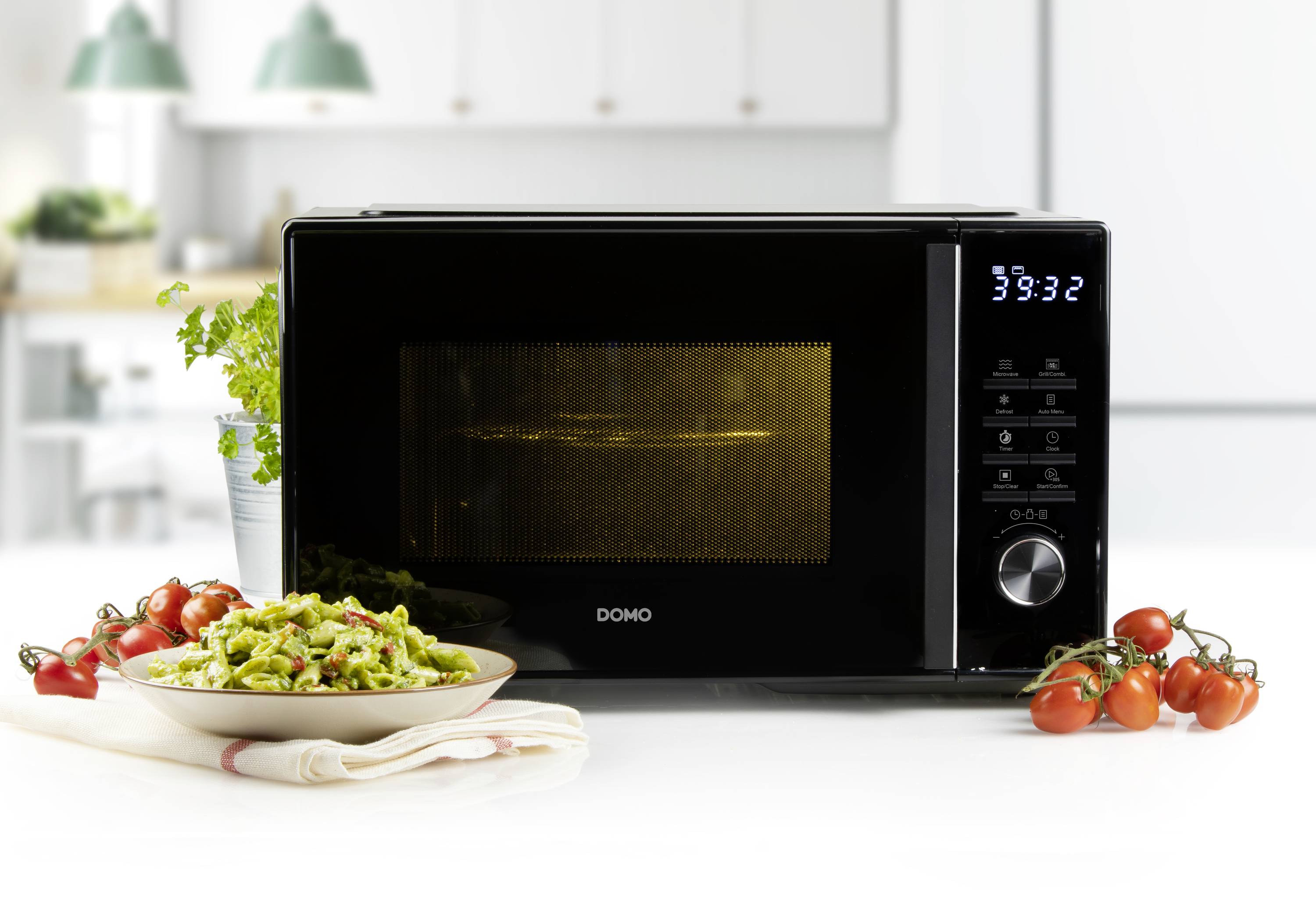 Un micro-ondes noir est posé sur une table blanche dans une cuisine. À côté se trouve une assiette de pâtes vertes accompagnées de tomates cerises.
