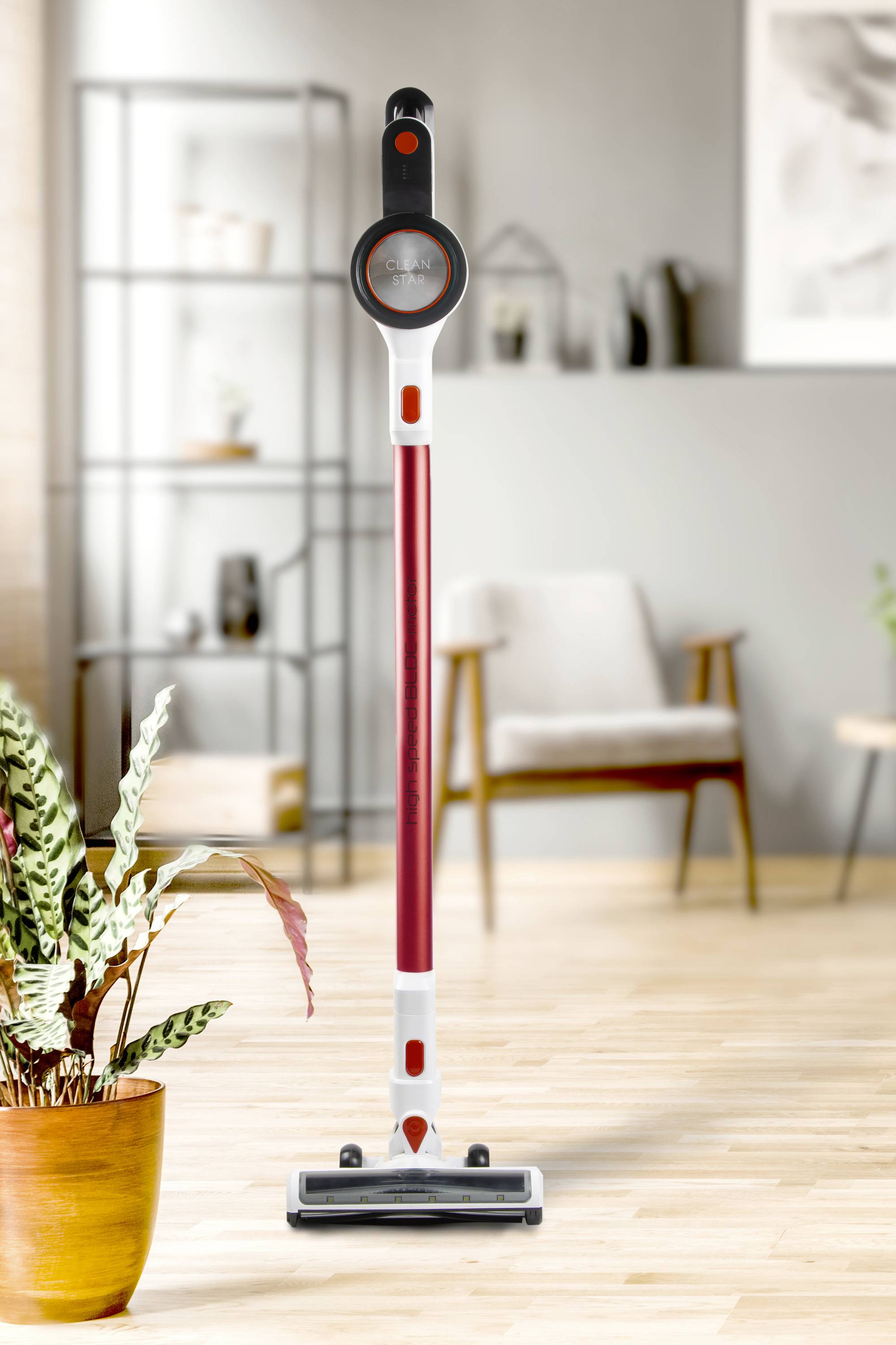 Un aspirateur sans fil rouge est debout dans un salon bien éclairé avec un sol en bois, des plantes et des meubles modernes.