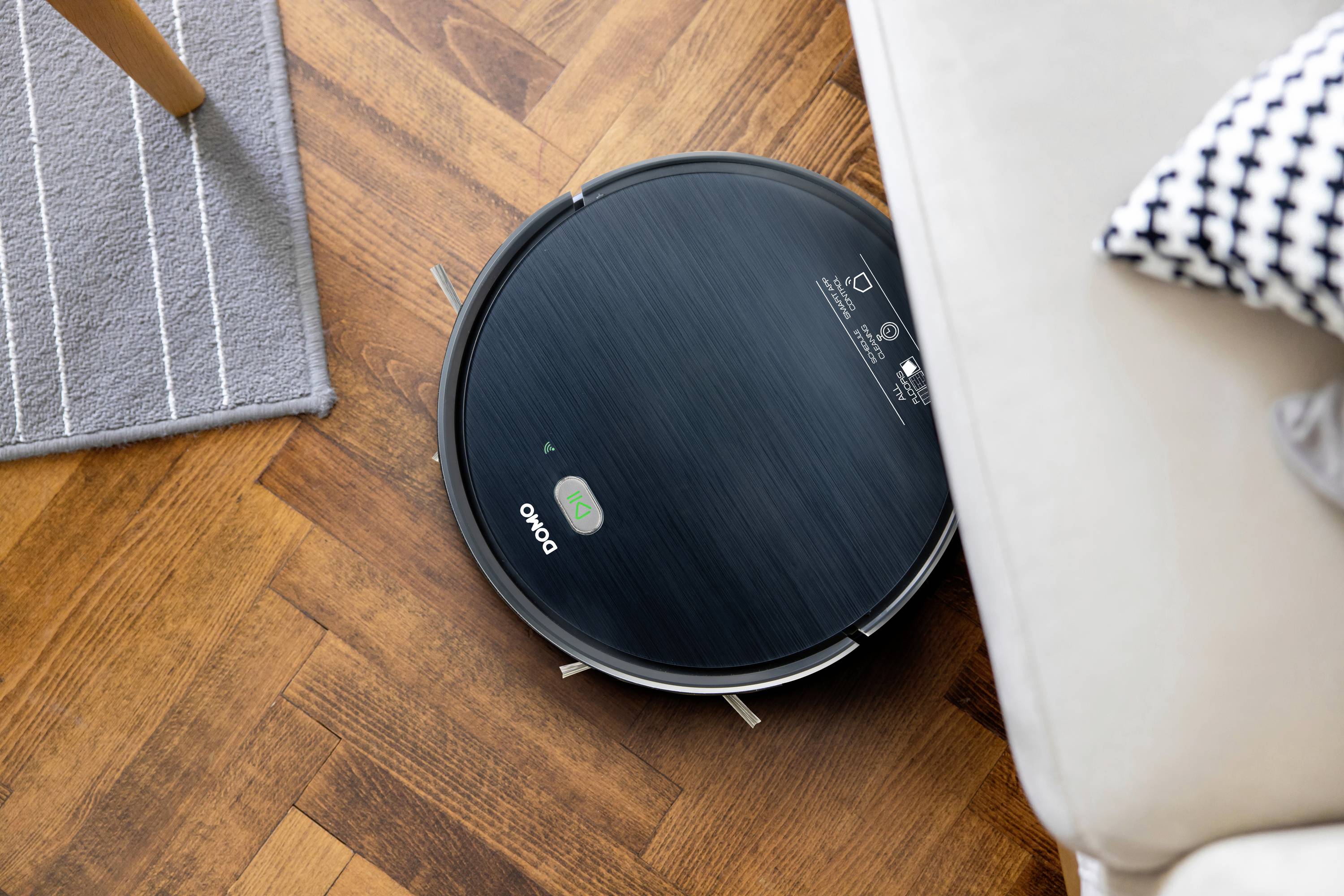 Un robot aspirateur rond nettoie un parquet près d'un tapis et d'un canapé.