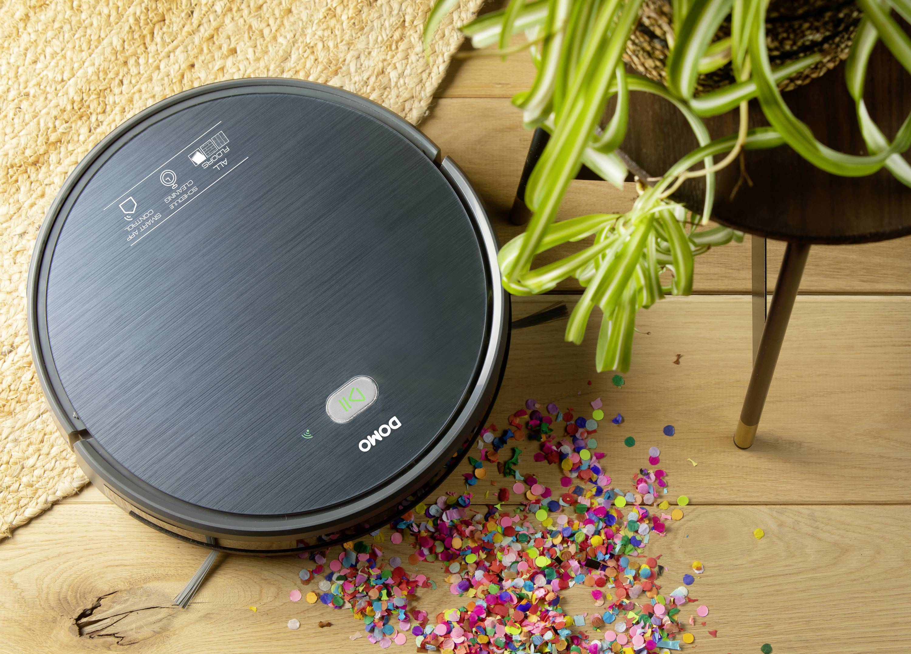 Un robot aspirateur aspire du confetti éparpillé sur un plancher en bois à côté d'une plante.