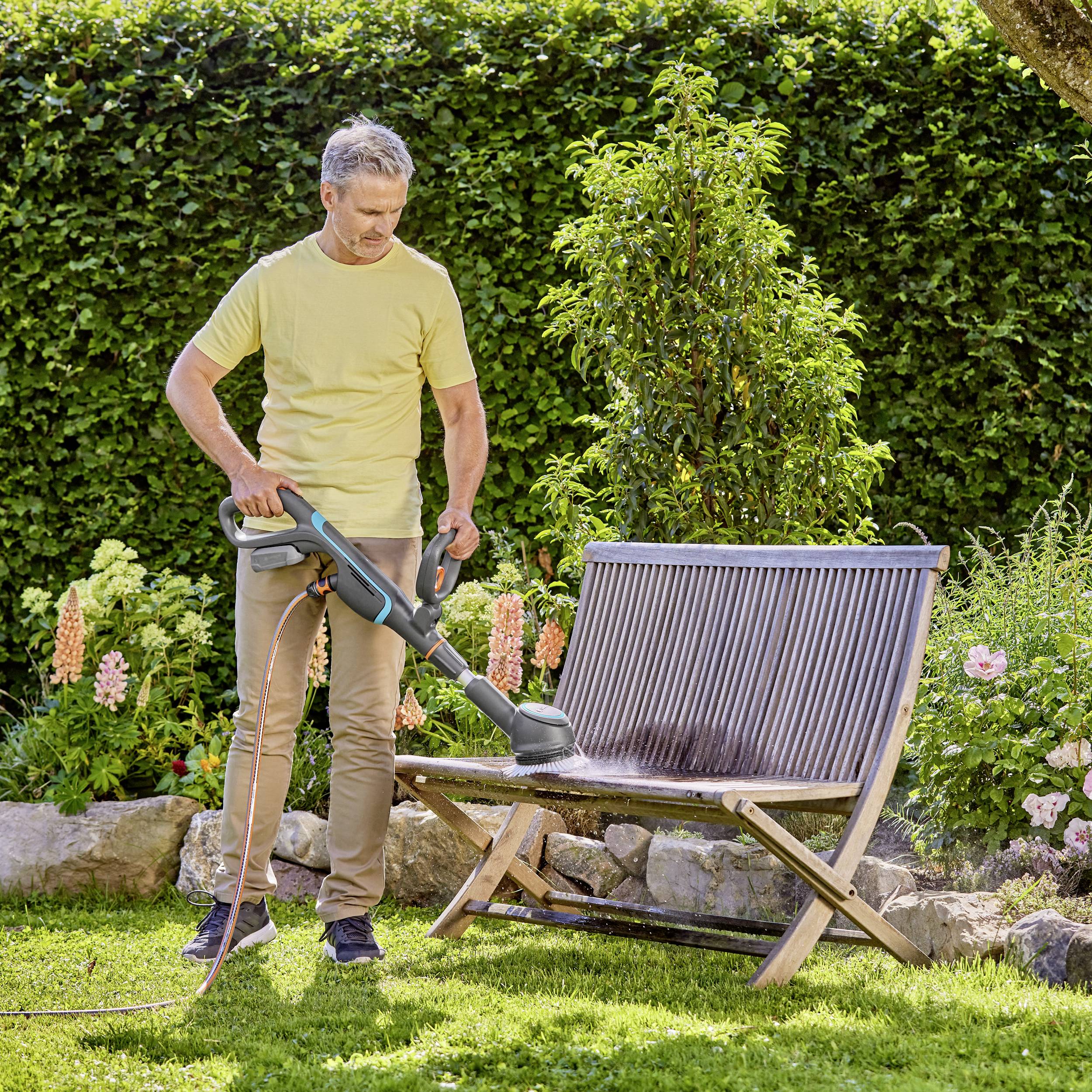 Un homme passe l'aspirateur sur un banc en bois dans le jardin, entouré de plantes et de pelouse, par une journée ensoleillée.